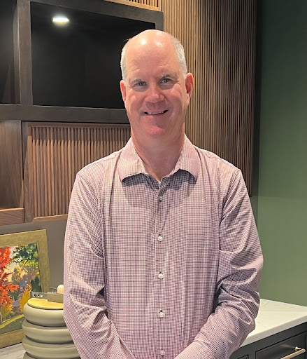 A man with short, light hair wearing a checked button-up shirt stands indoors, smiling, surrounded by art and decor—reflecting his expertise in luxury interior design as an interior designer in Massachusetts.