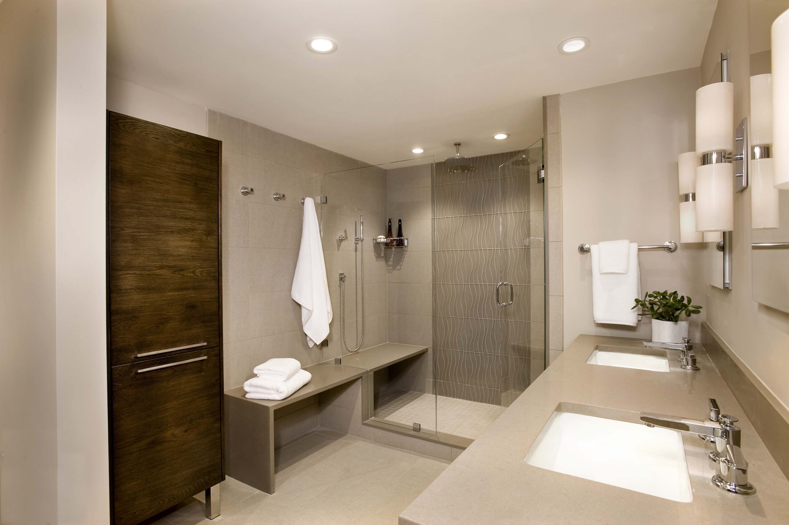 Modern bathroom by an Interior Designer in Massachusetts, featuring a glass-enclosed walk-in shower, double sinks, a wooden cabinet, wall-mounted lights, and neatly folded towels for a touch of luxury interior design.