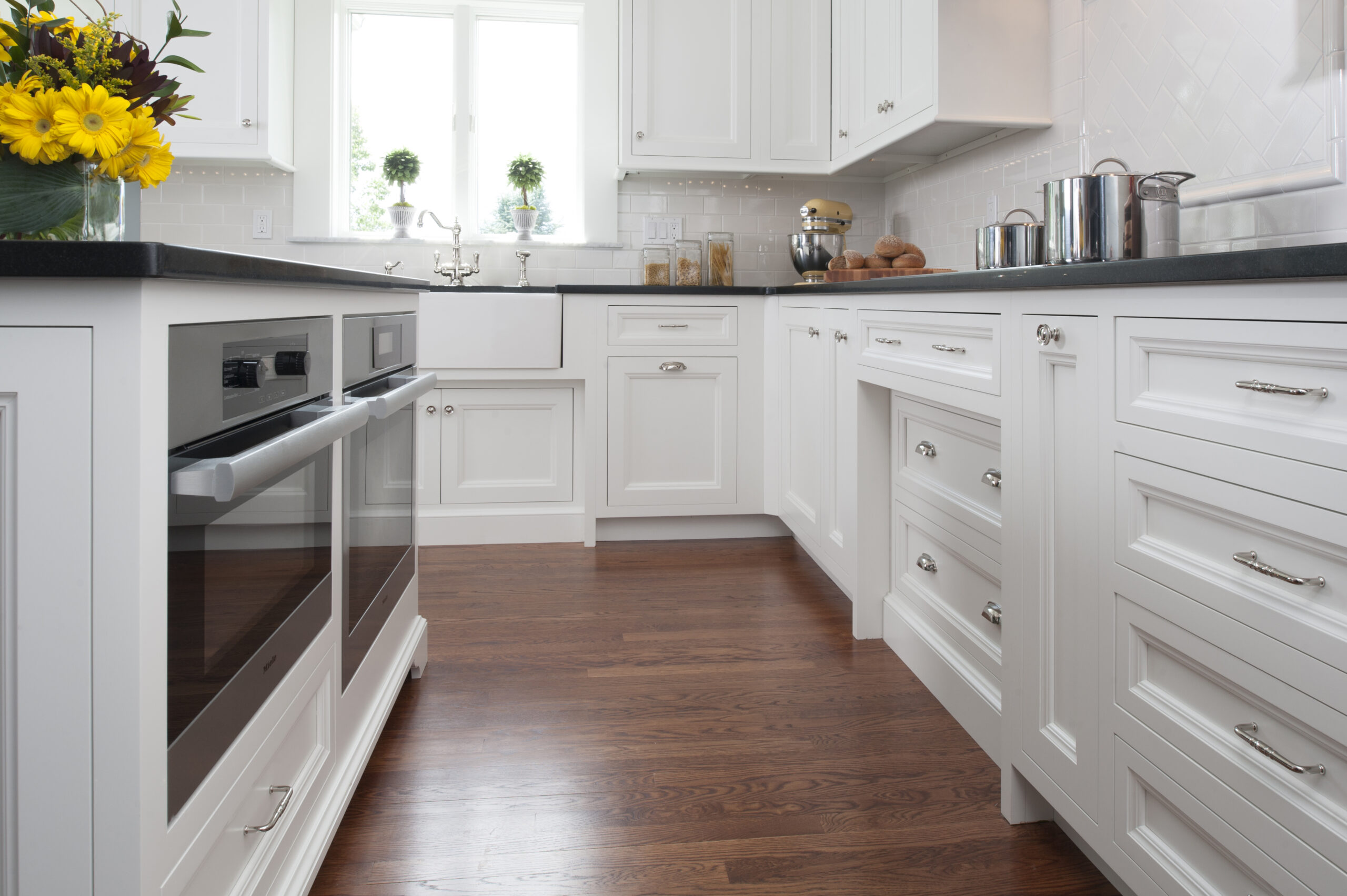 Modern kitchen with white cabinets, dark countertops, stainless steel appliances, a bouquet of yellow flowers, and wooden flooring, featuring sunlight streaming in—an elegant example of luxury interior design.