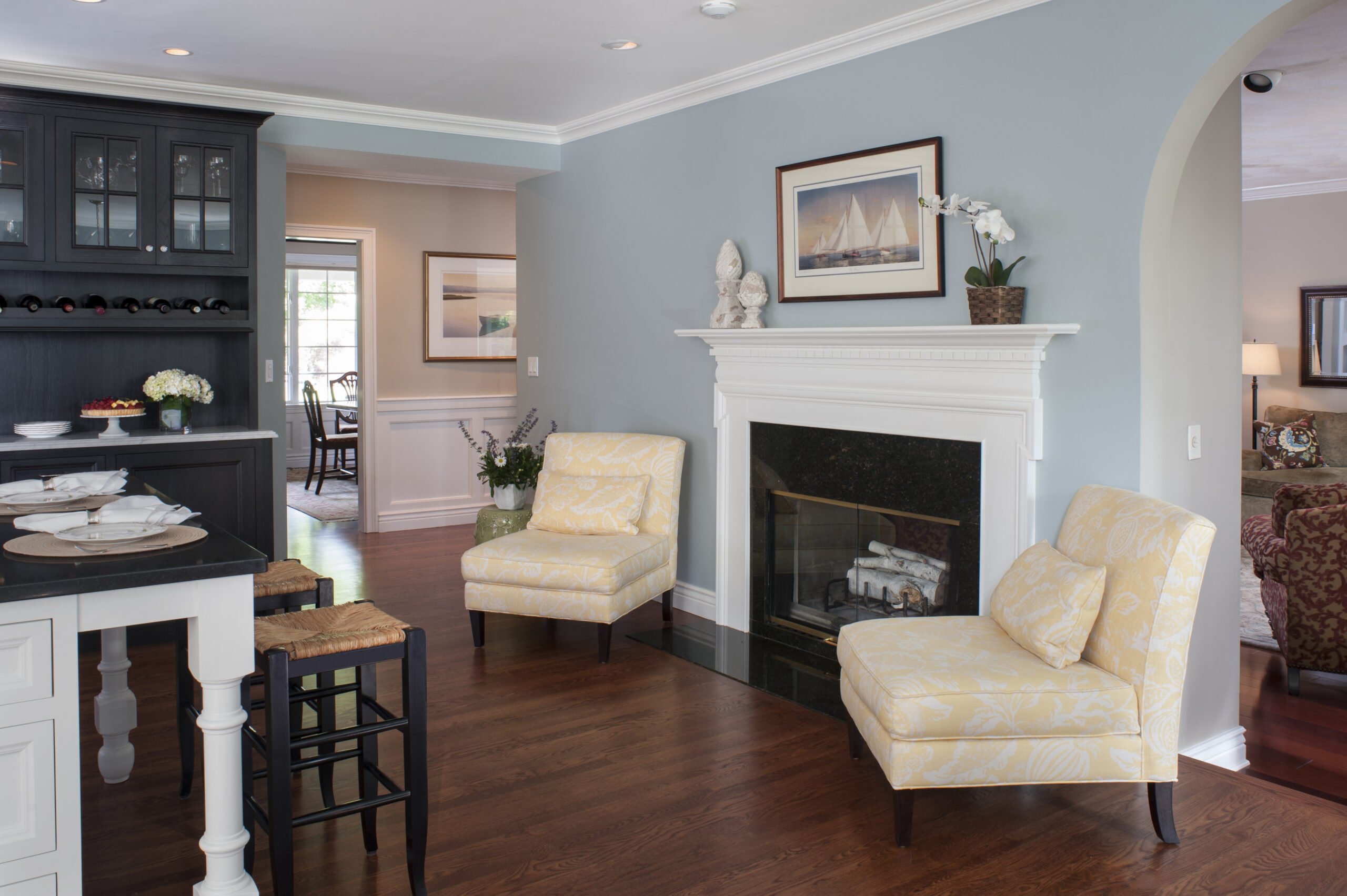 A living area with two yellow patterned chairs near a white fireplace, hardwood floors, a kitchen island with stools, and wall art in a bright, open room by a leading Interior Designer Massachusetts specializing in Luxury Interior Design.