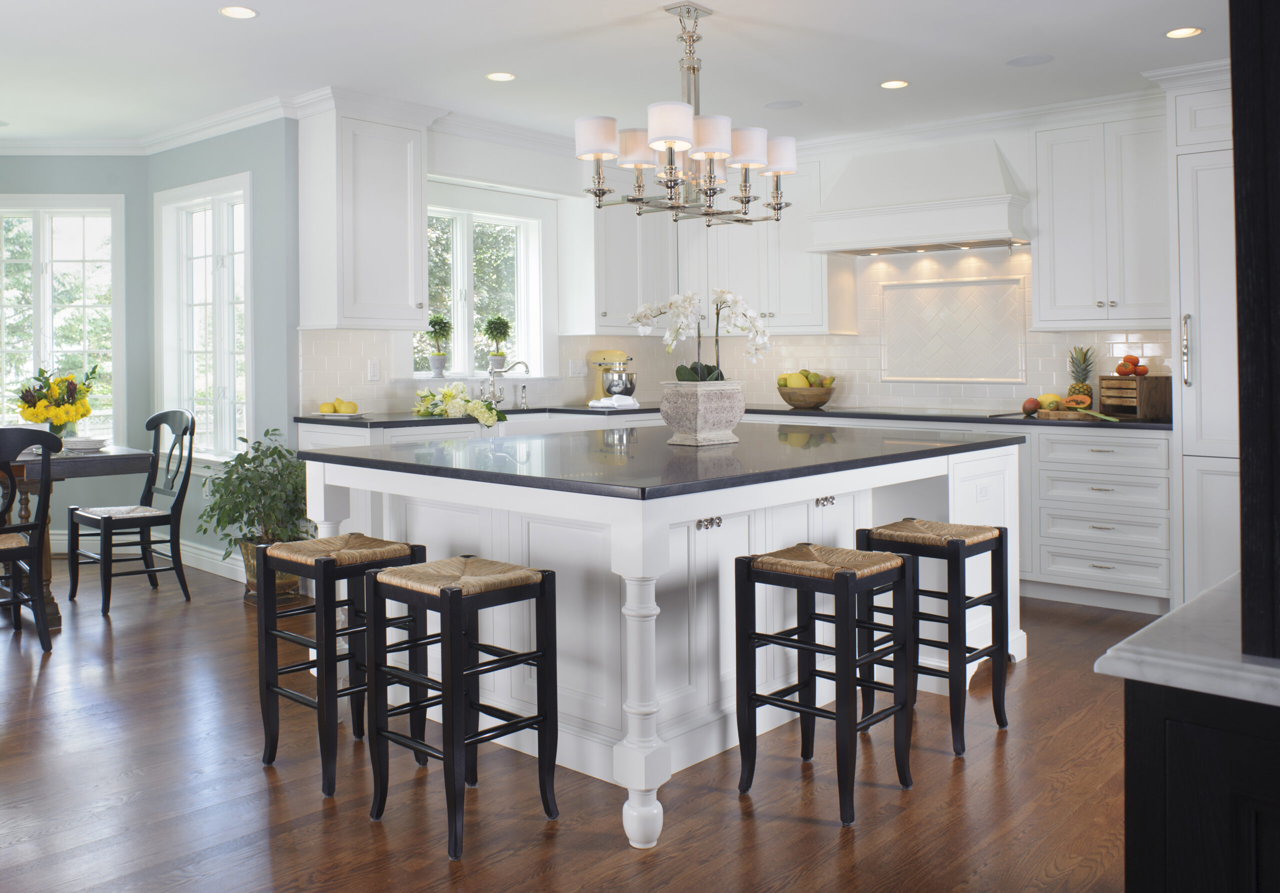 Bright, modern kitchen with a large white island, black countertop, four stools, hardwood floors, and white cabinetry. A chandelier hangs above the island—perfect for those who appreciate Luxury Interior Design.