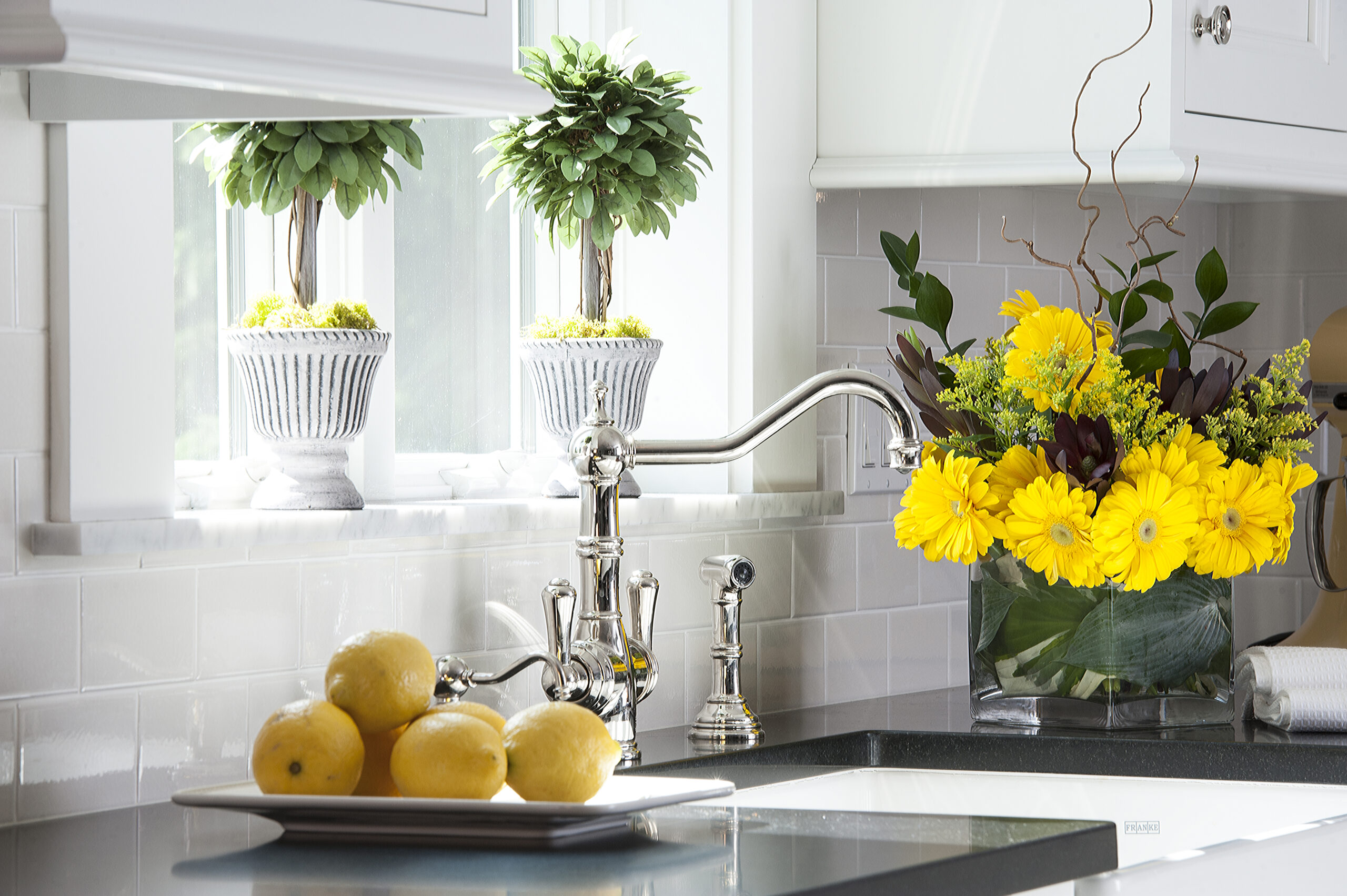 A kitchen sink with a chrome faucet, potted plants on the windowsill, a bouquet of yellow flowers, and a plate of lemons on the countertop showcases luxury interior design at its finest.