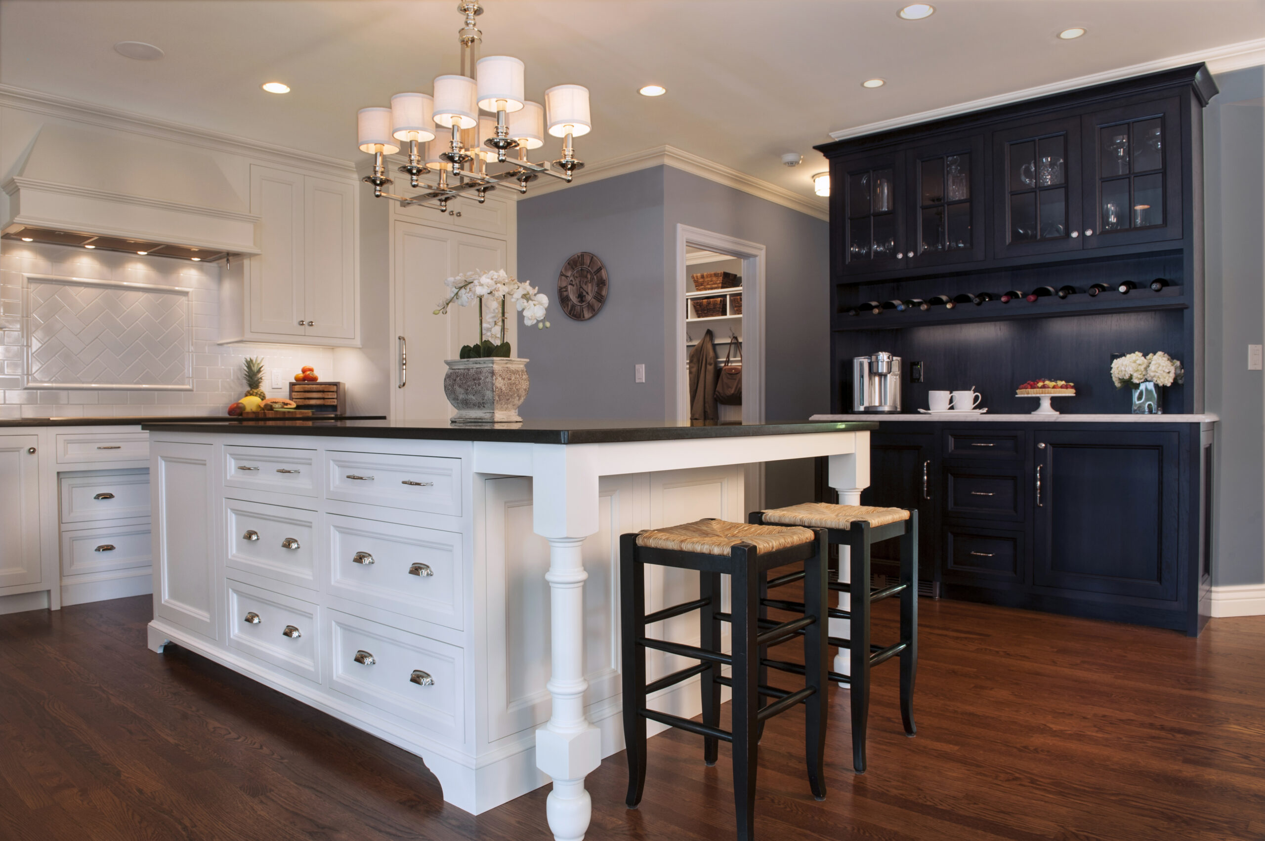 A modern kitchen with white cabinets, a large island with stools, dark wood floors, a black beverage cabinet, and a chandelier overhead showcases luxury interior design by an acclaimed interior designer in Massachusetts.