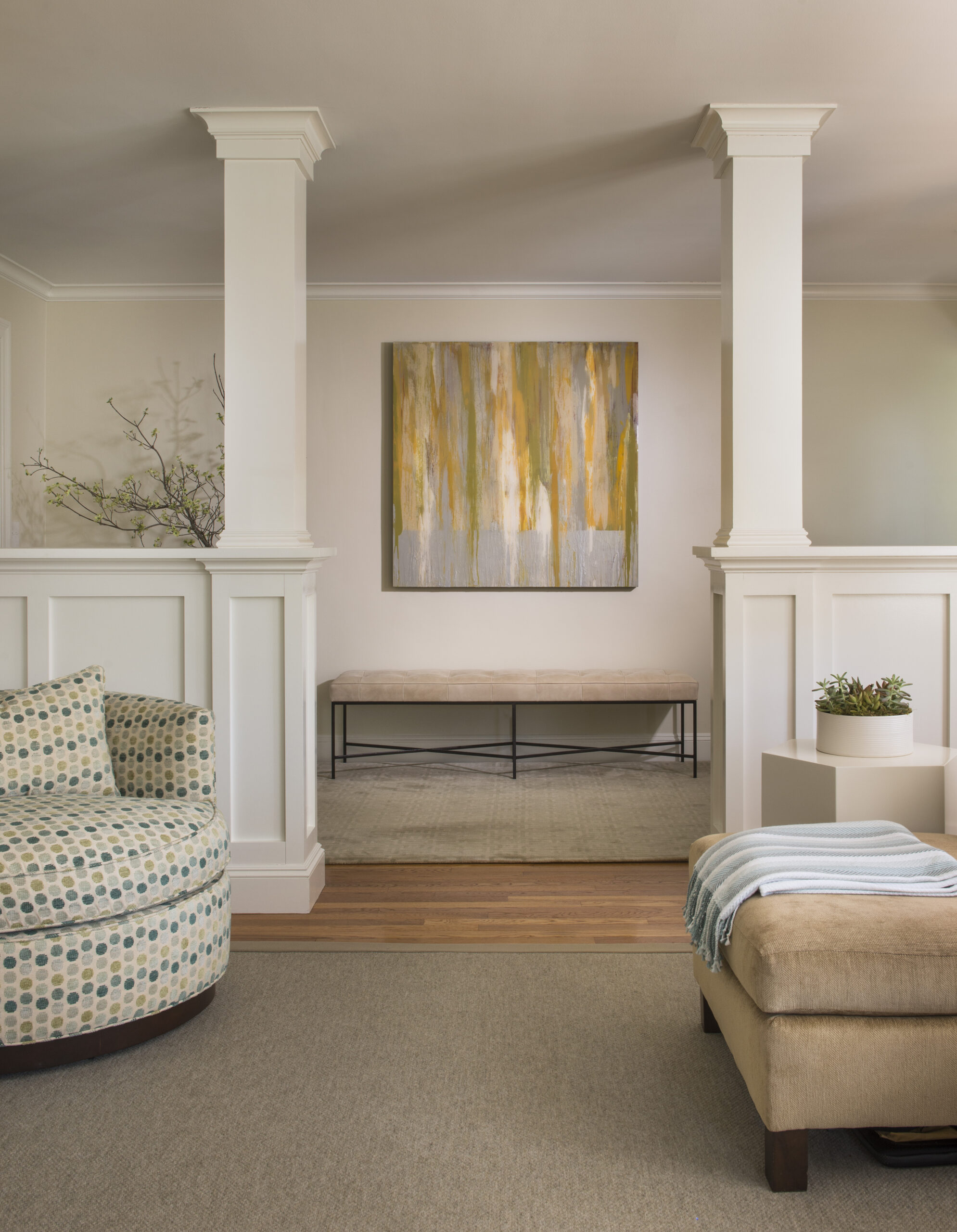 A modern living room by a leading Interior Designer Massachusetts features two white columns, abstract yellow and gray artwork, a patterned chair, beige bench, and a small plant—showcasing luxury interior design at its finest.