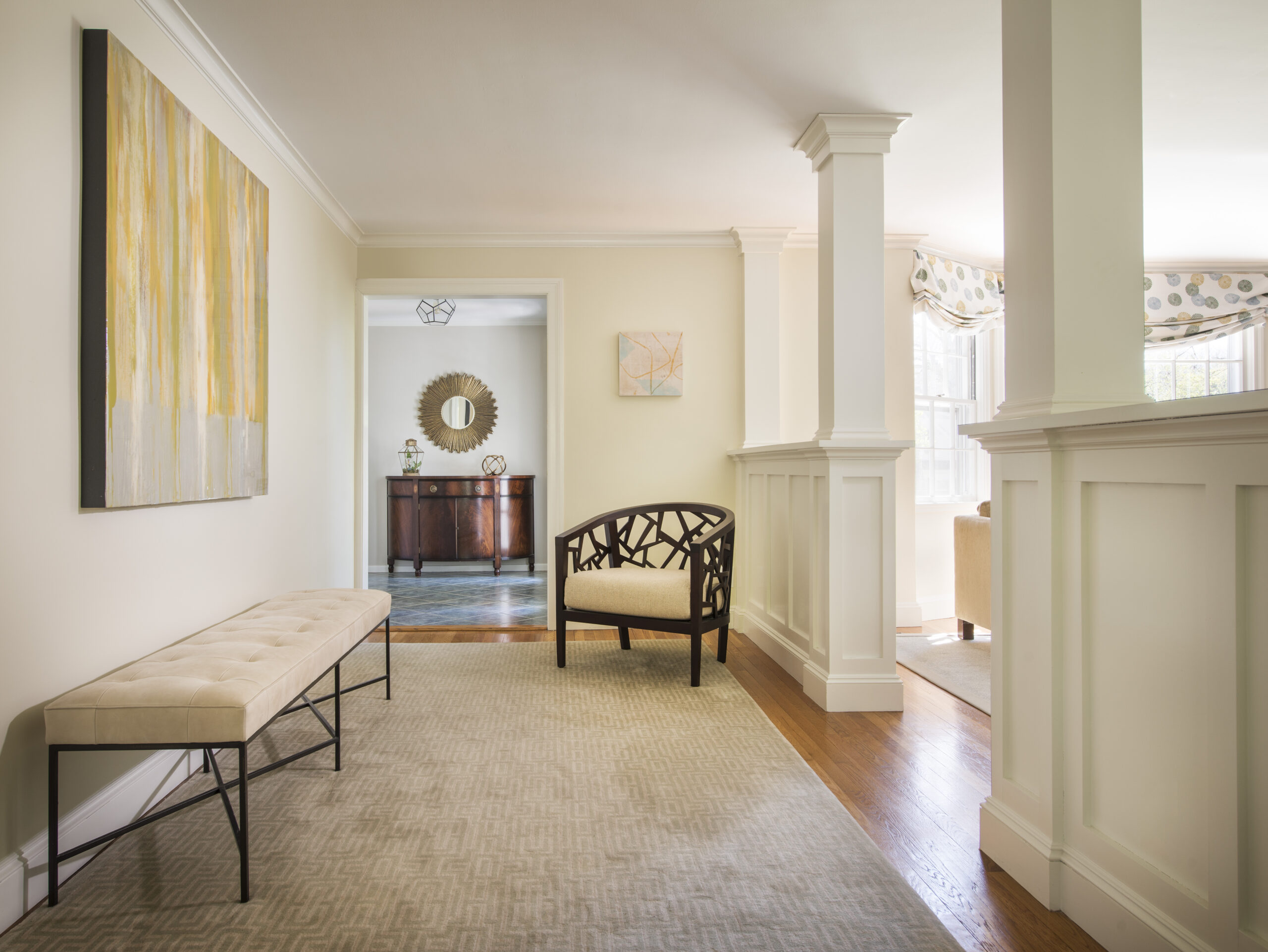 A hallway curated with luxury interior design features a beige bench, abstract wall art, a patterned chair, and a view into a room with a console table, circular mirror, and hardwood floors—showcasing the touch of an expert interior designer Massachusetts.
