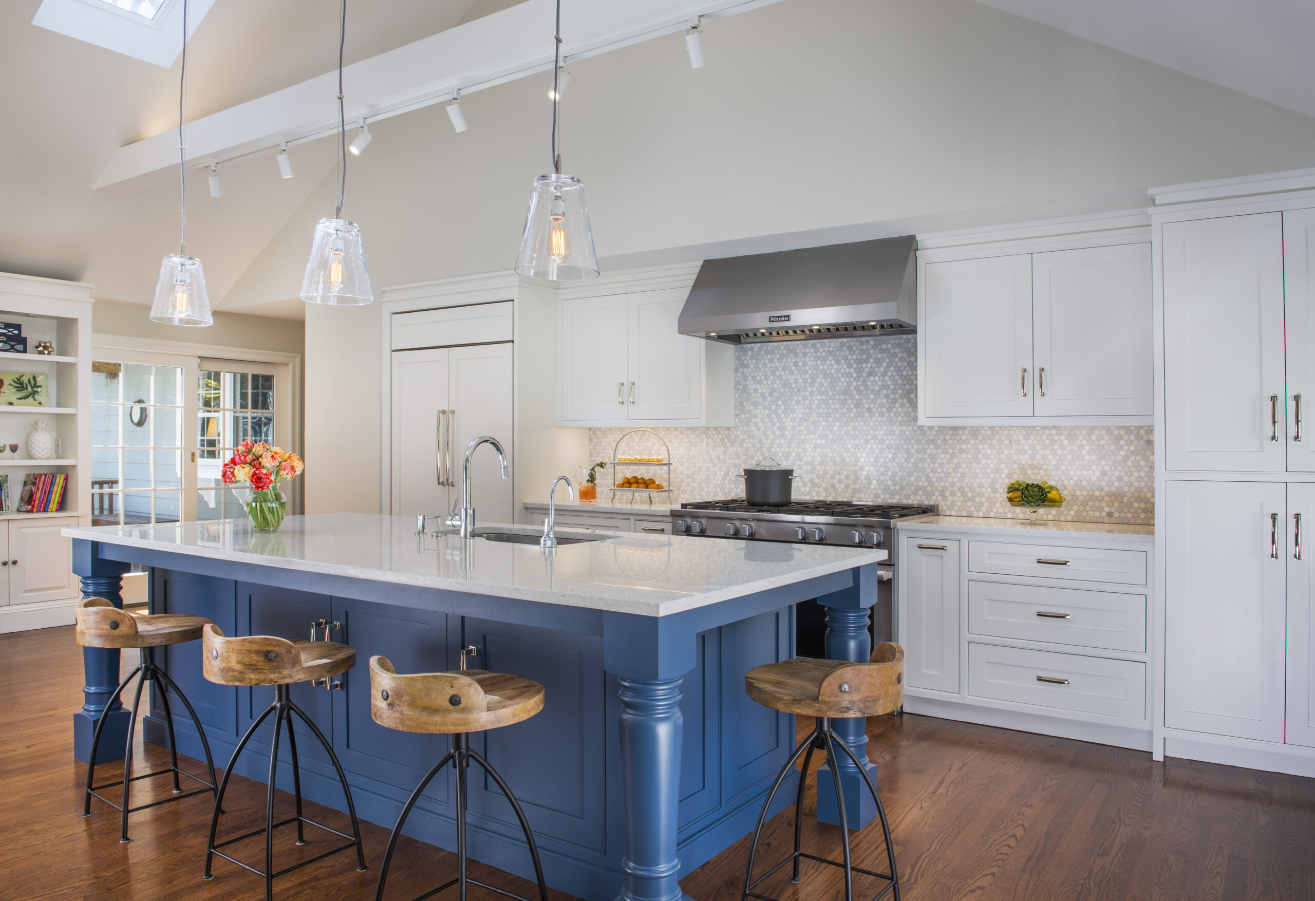 Modern kitchen with a blue island, wooden stools, white cabinets, pendant lights, and stainless steel appliances on a hardwood floor. A vase of flowers sits on the island—an example of luxury interior design by an Interior Designer in Massachusetts.