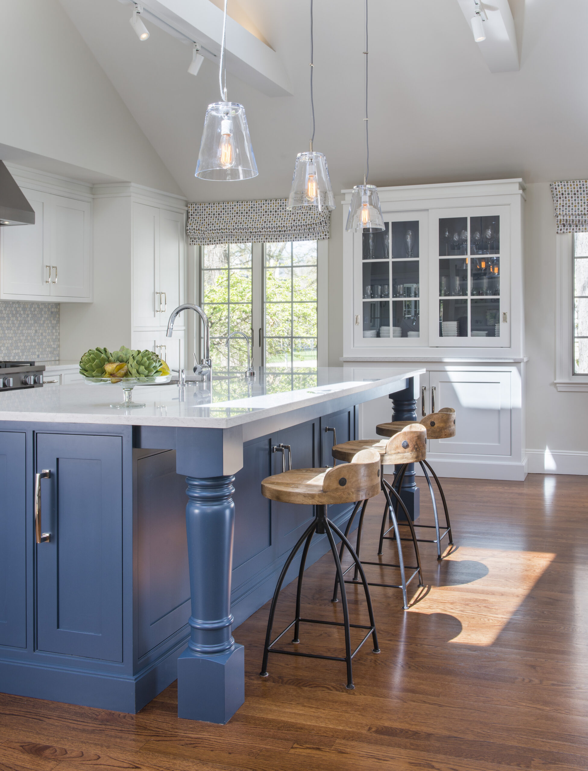Modern kitchen with a large island featuring blue cabinetry, wooden stools, pendant lights, and white cabinets. Large windows fill the space with light—an example of Luxury Interior Design by an expert Interior Designer Massachusetts.