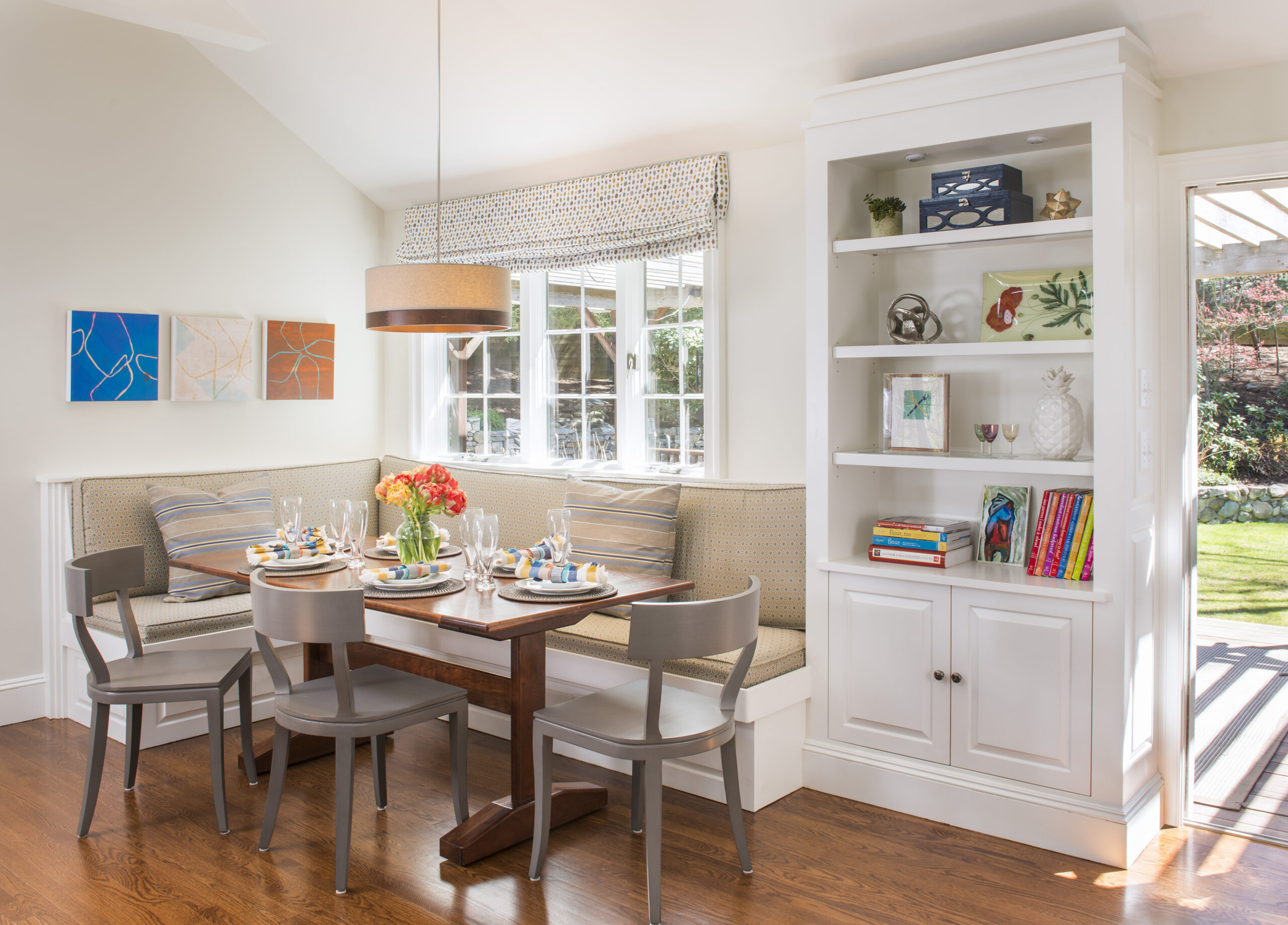 A bright dining nook designed by an Interior Designer Massachusetts, featuring a wooden table, four chairs, built-in bench seating, a pendant light, wall art, and a white bookshelf filled with decor and books.