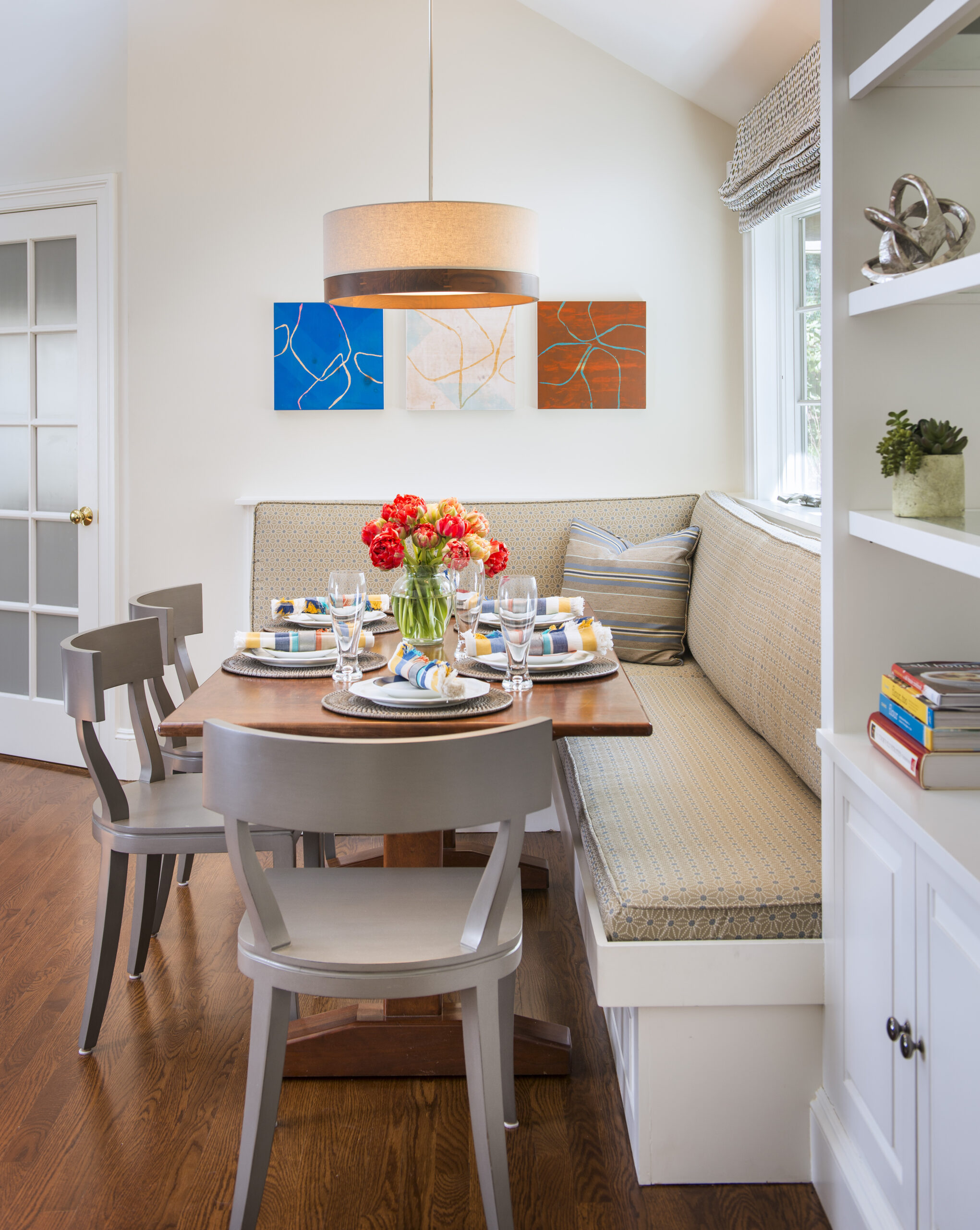 A dining nook designed by an Interior Designer Massachusetts features a wooden table, four gray chairs, a built-in bench, set tableware, red flowers, wall art, and shelves with books and decor on hardwood floors.