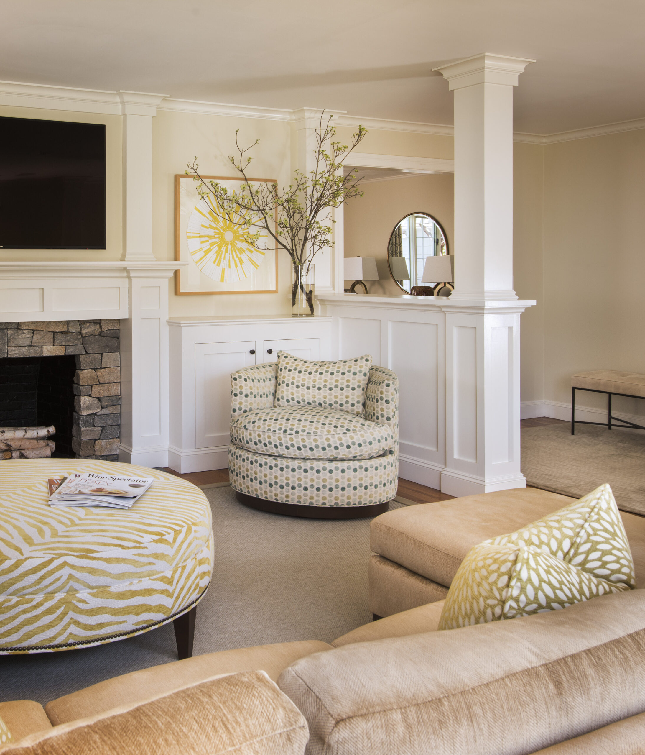 Light-filled living room with neutral furnishings, patterned ottoman and chair, fireplace with stone details, wall art, and a vase with branches on a white partition—a perfect example of luxury interior design.