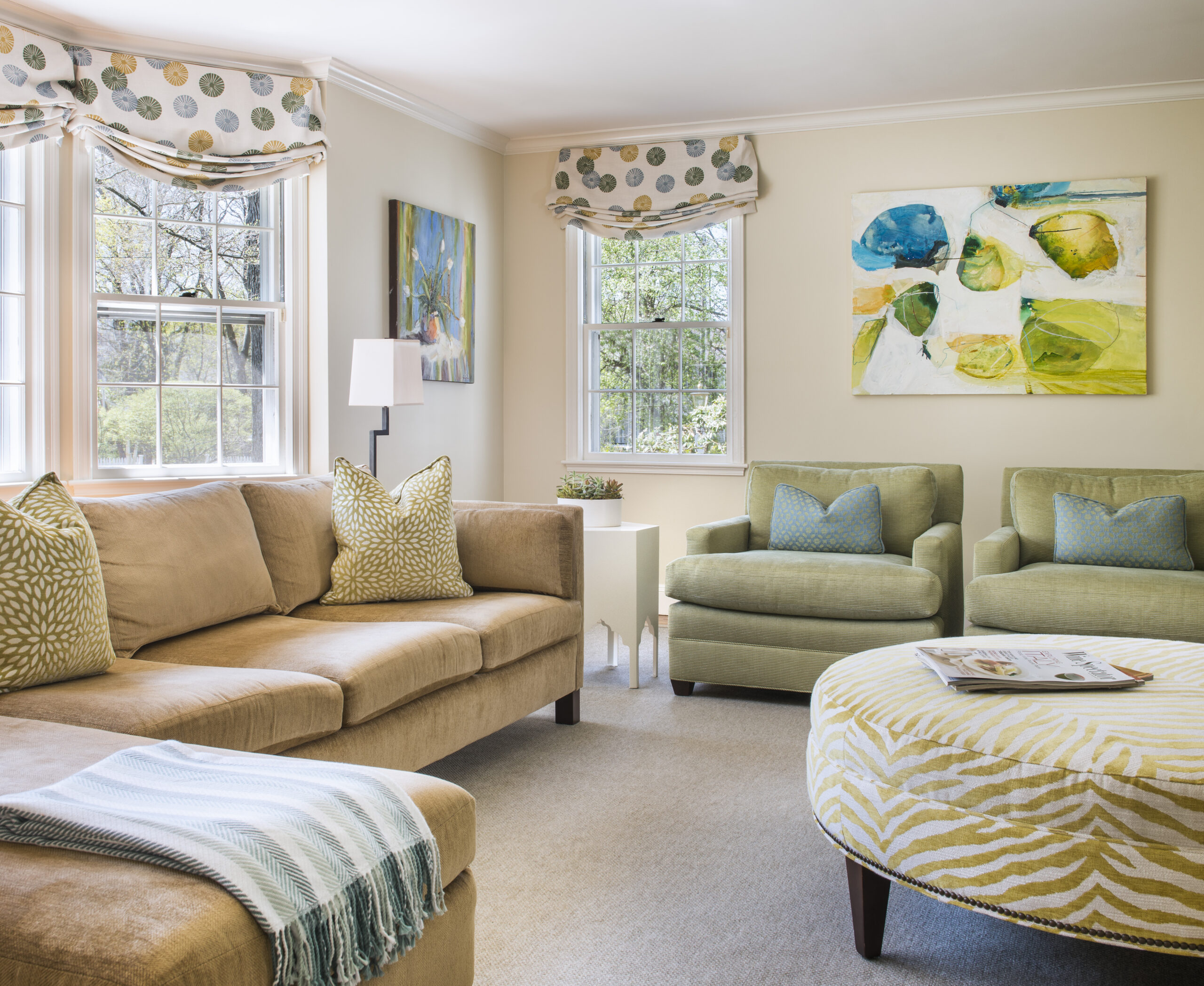 Bright living room with beige sofa, two green armchairs, patterned ottoman, abstract wall art, and large windows with valances letting in natural light—perfectly styled for luxury interior design by a top Interior Designer Massachusetts.