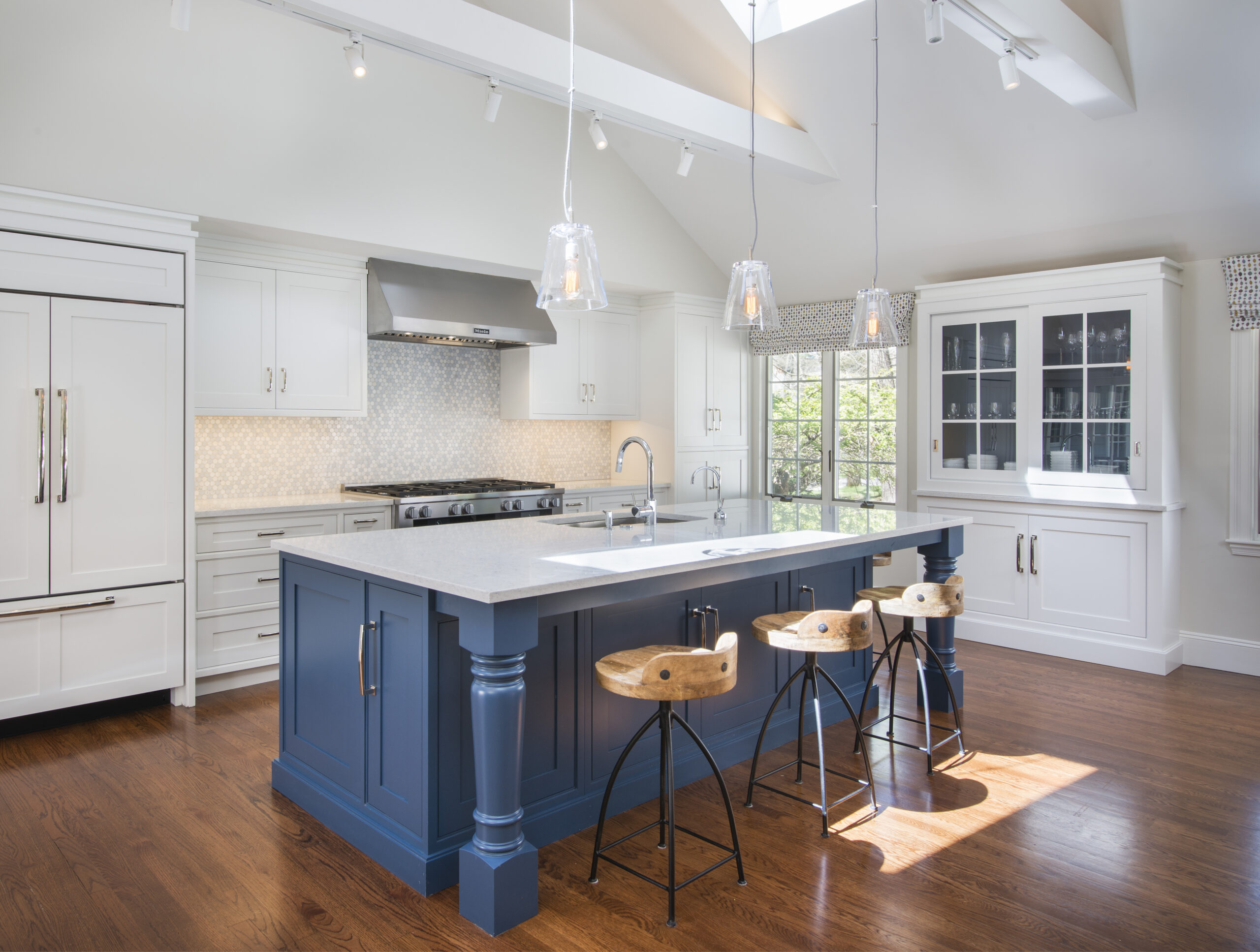 Modern kitchen with white cabinets, a blue island and wooden stools, pendant lights, stainless steel appliances, and hardwood flooring—sunlight streams through a large window. Perfect for those seeking luxury interior design by an expert interior designer in Massachusetts.