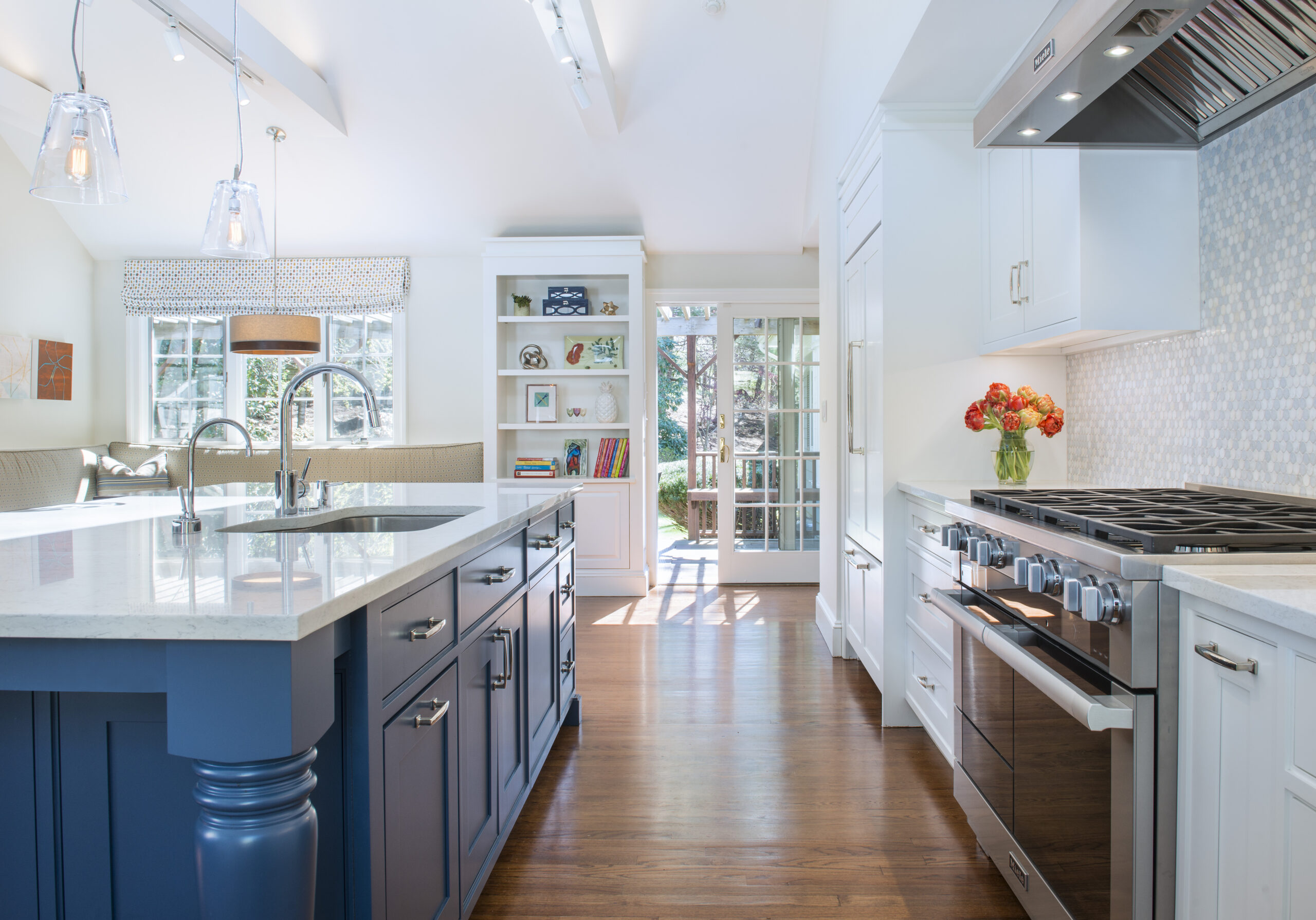 Bright, modern kitchen showcasing luxury interior design, with a blue island, white cabinets, stainless steel appliances, a vase of orange flowers, and sunlight streaming through glass doors.