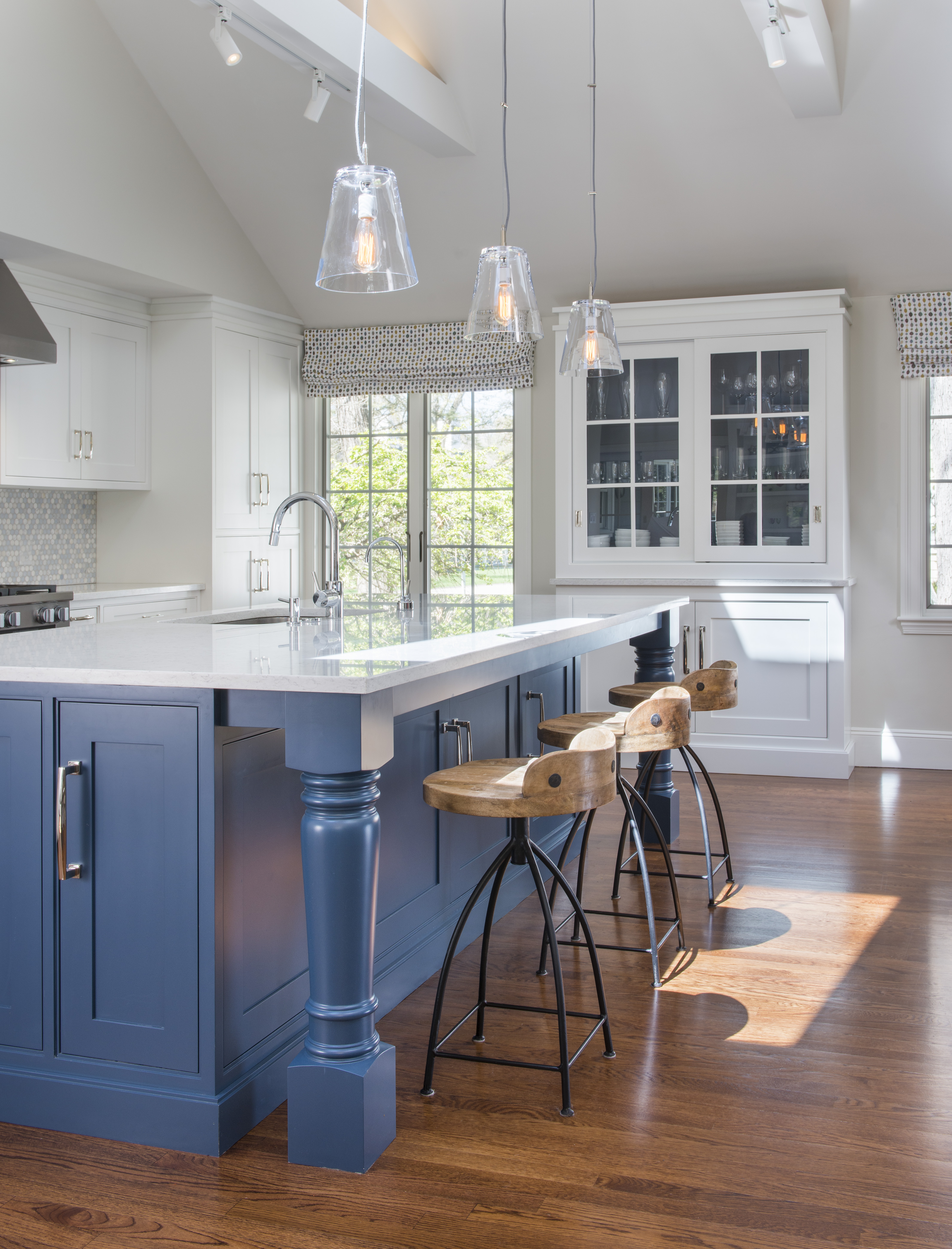 Modern kitchen with a blue island, wooden stools, white cabinets, pendant lights, and an open sightline to the family room, designed by an interior designer in Massachusetts.