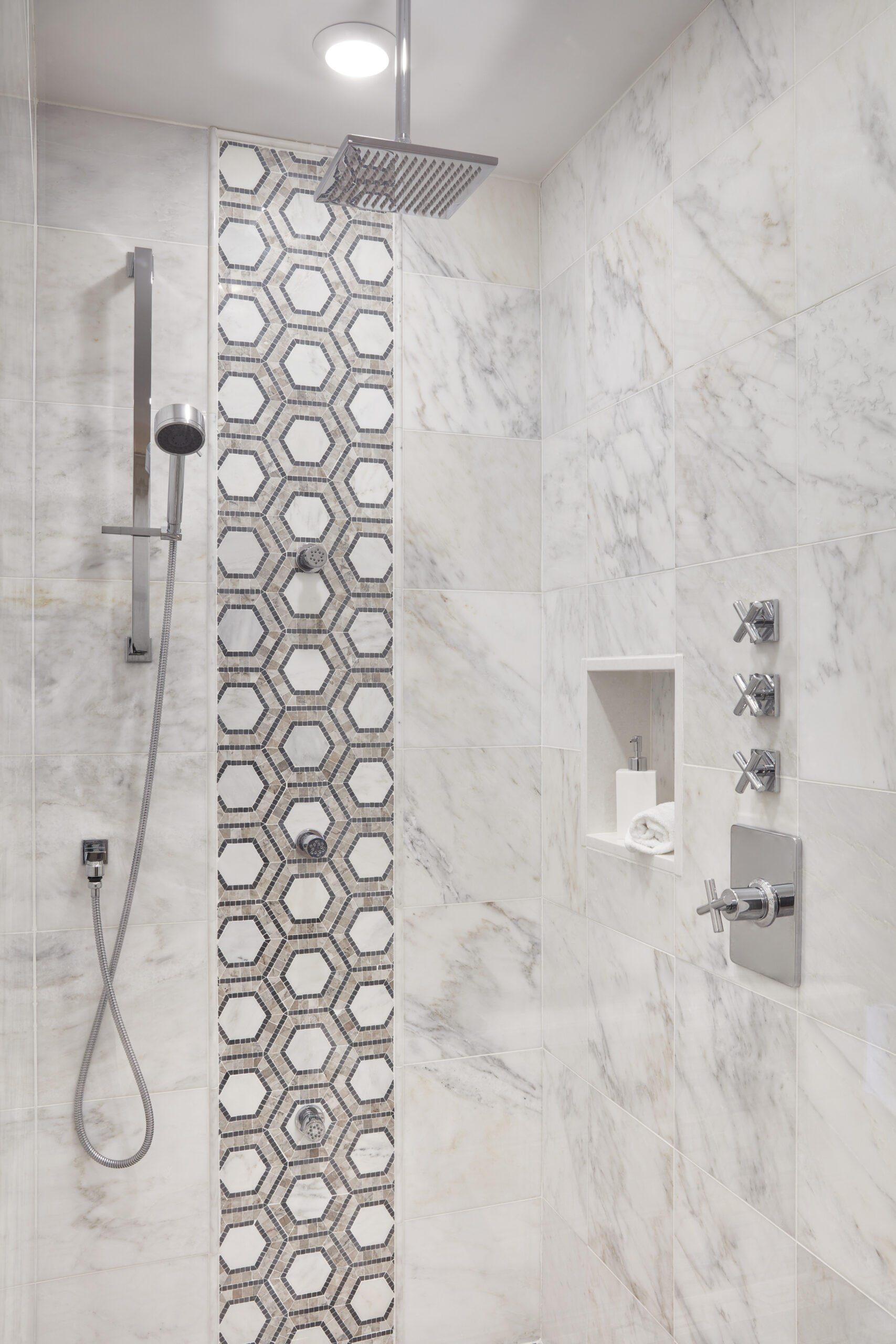 Modern shower with marble walls, hexagonal mosaic tile accent, built-in niche with soap, rainfall and handheld showerheads, and multiple control knobs—a perfect example of Luxury Interior Design by an expert Interior Designer in Massachusetts.