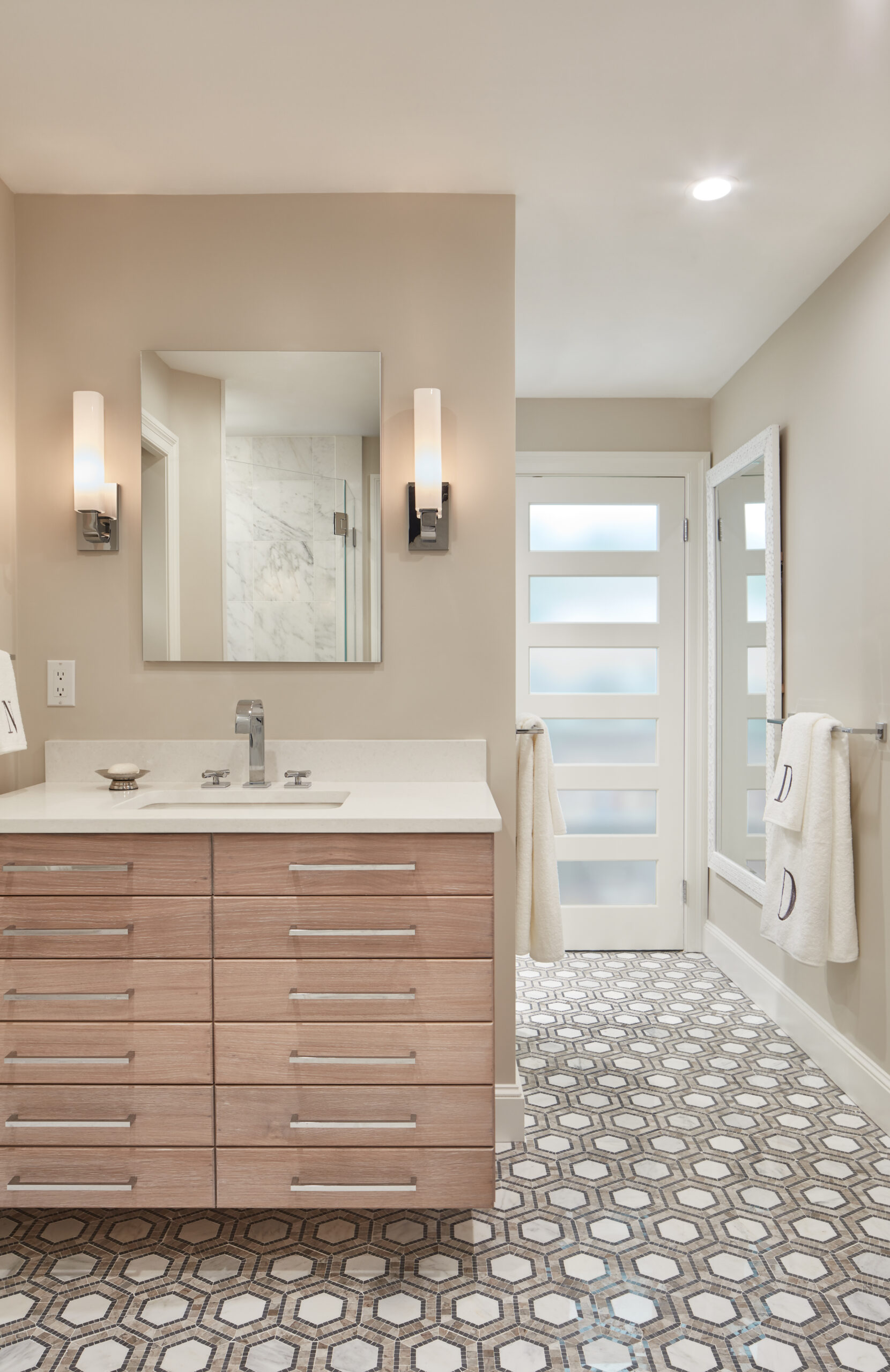 Modern bathroom with light wood vanity, rectangular mirror, wall sconces, hexagonal tile floor, and frosted glass door—an example of luxury interior design by an expert interior designer in Massachusetts. Towels hang neatly on racks.
