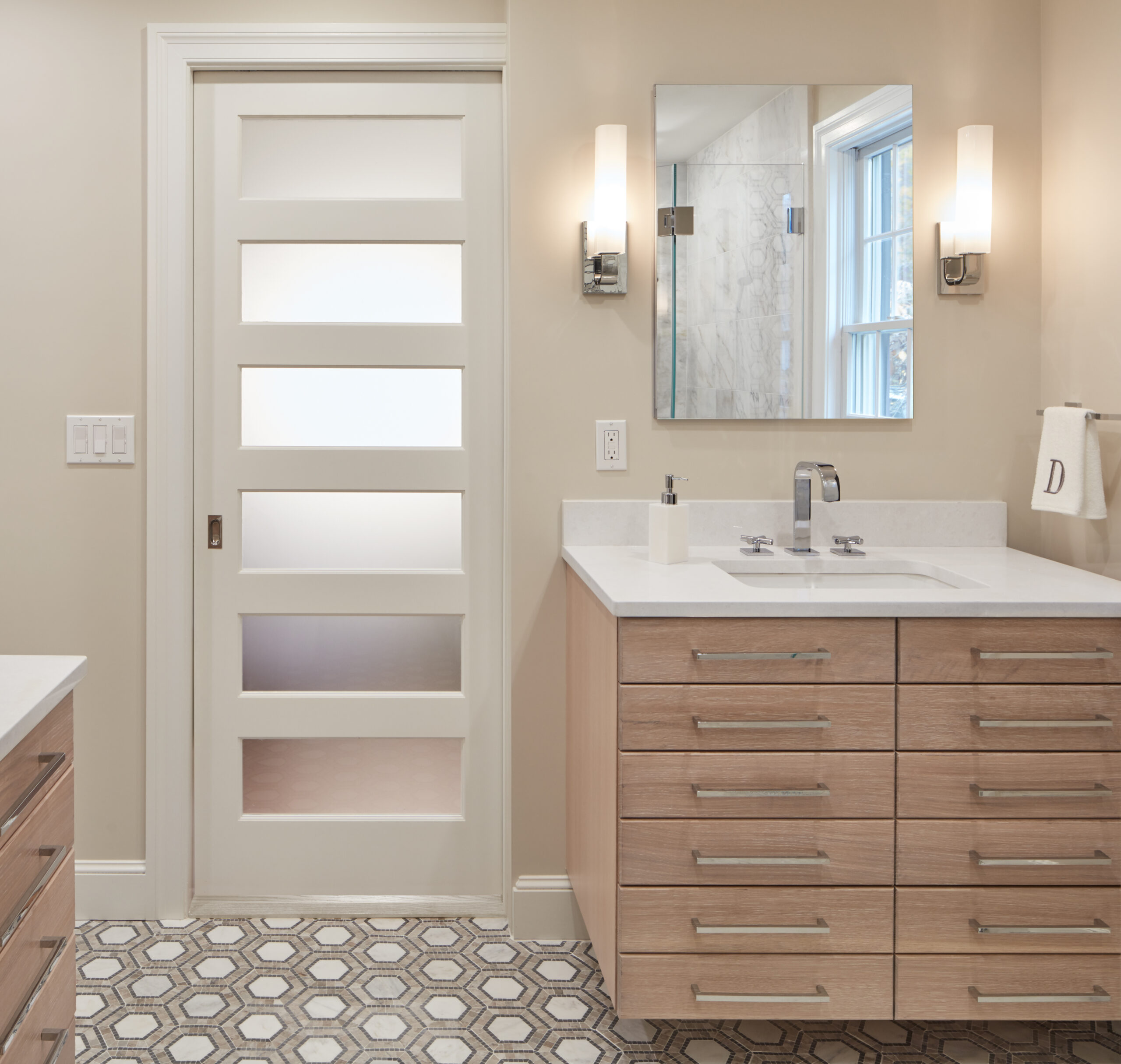 Modern bathroom with light wood vanity, rectangular mirror, wall sconces, geometric tile floor, and a white sliding door with frosted glass panels—a stunning example of luxury interior design by an interior designer Massachusetts trusts.