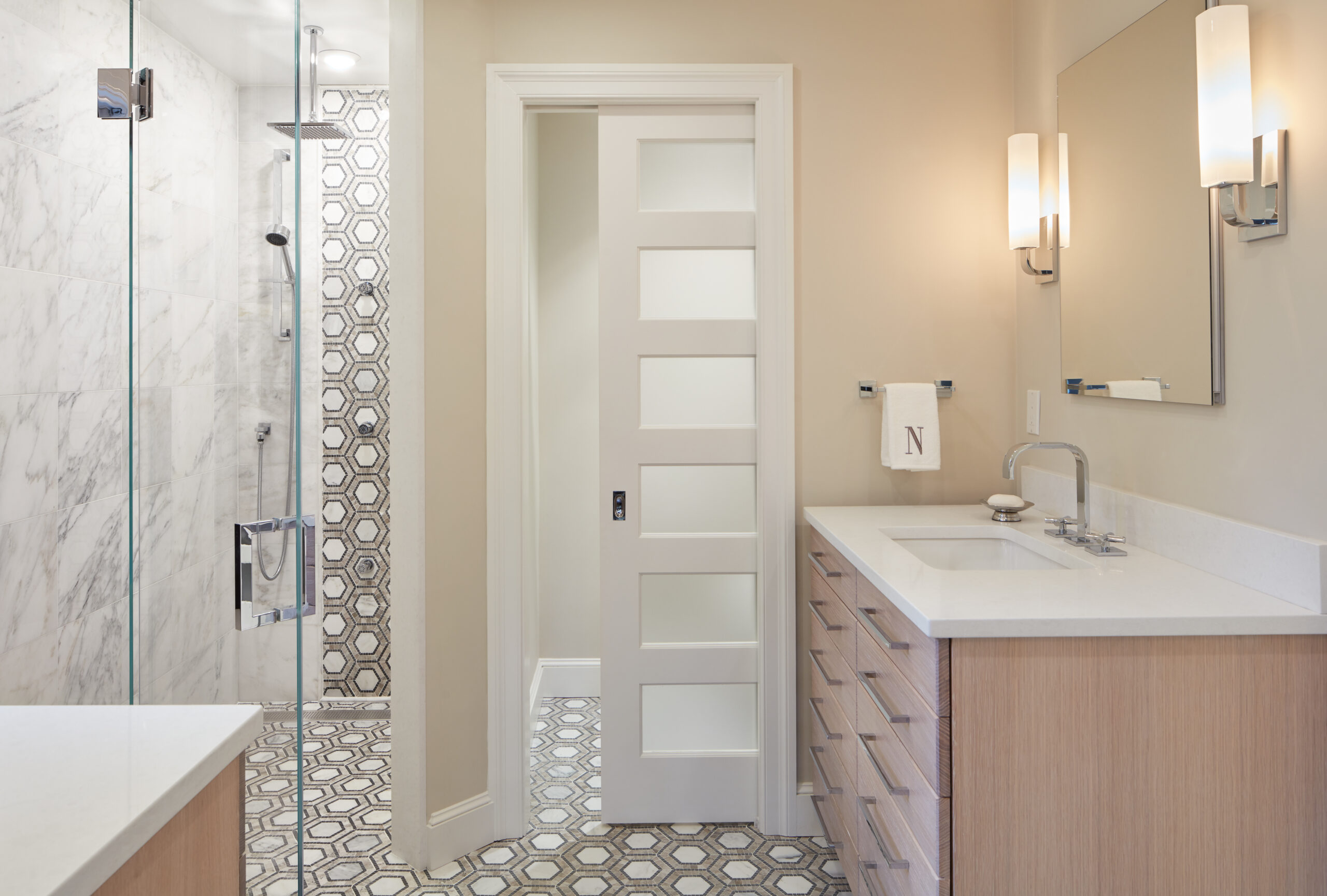 Modern bathroom with a sliding door, light wood vanity, rectangular sink, wall-mounted lights, and a walk-in shower featuring marble walls and hexagonal tile floor—showcasing luxury interior design by an expert interior designer in Massachusetts.