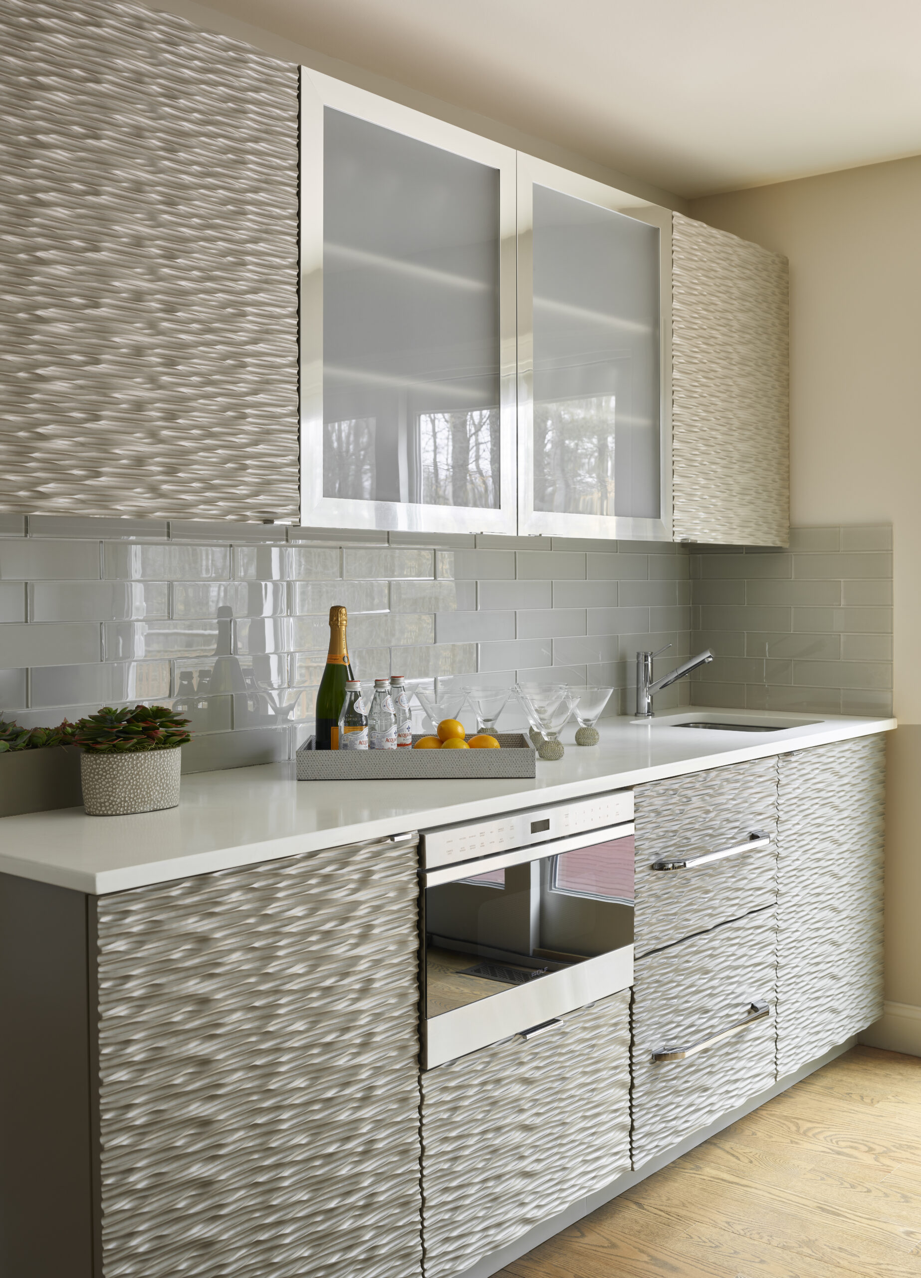 Modern kitchen with textured cabinets, glass-front upper cupboards, gray subway tile backsplash, built-in appliances, and a countertop sink—styled by a Luxury Interior Design expert—with drinks set out alongside glasses, lemons, and a potted plant.