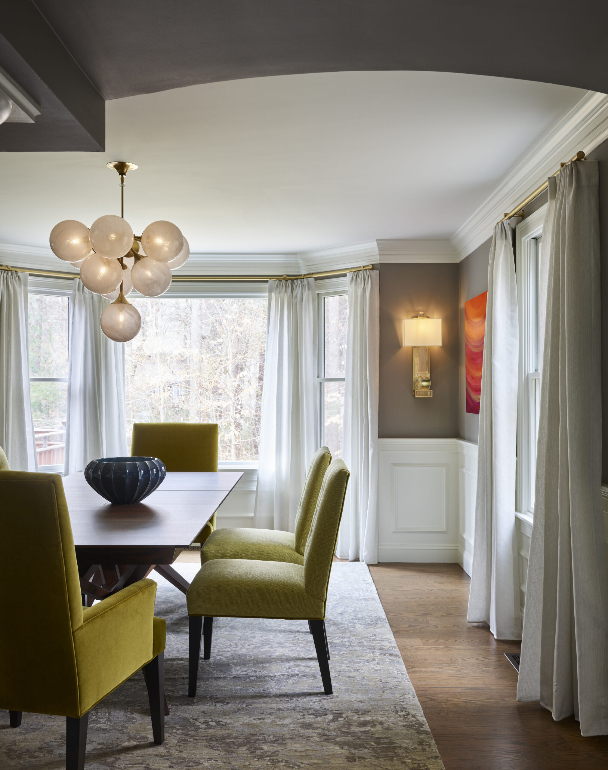 A modern dining room with a wooden table, yellow upholstered chairs, a cluster globe chandelier, and sheer white curtains showcases the refined touch of an Interior Designer Massachusetts, offering luxury interior design with a serene view of trees.