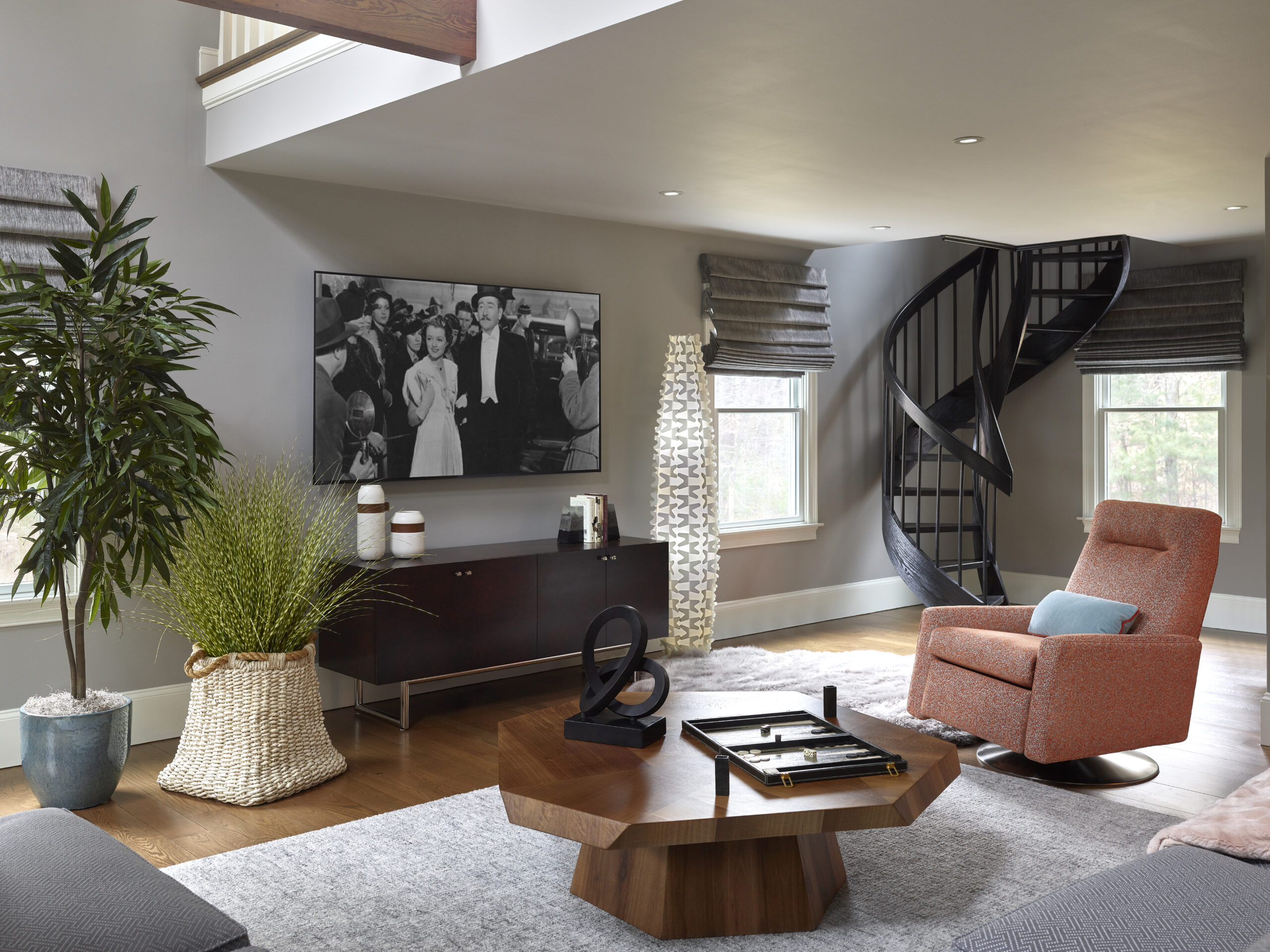 Modern living room with a spiral staircase, orange swivel chair, large plant, geometric wood coffee table, and TV displaying a black-and-white movie. Natural light streams in—perfect inspiration for luxury interior design.