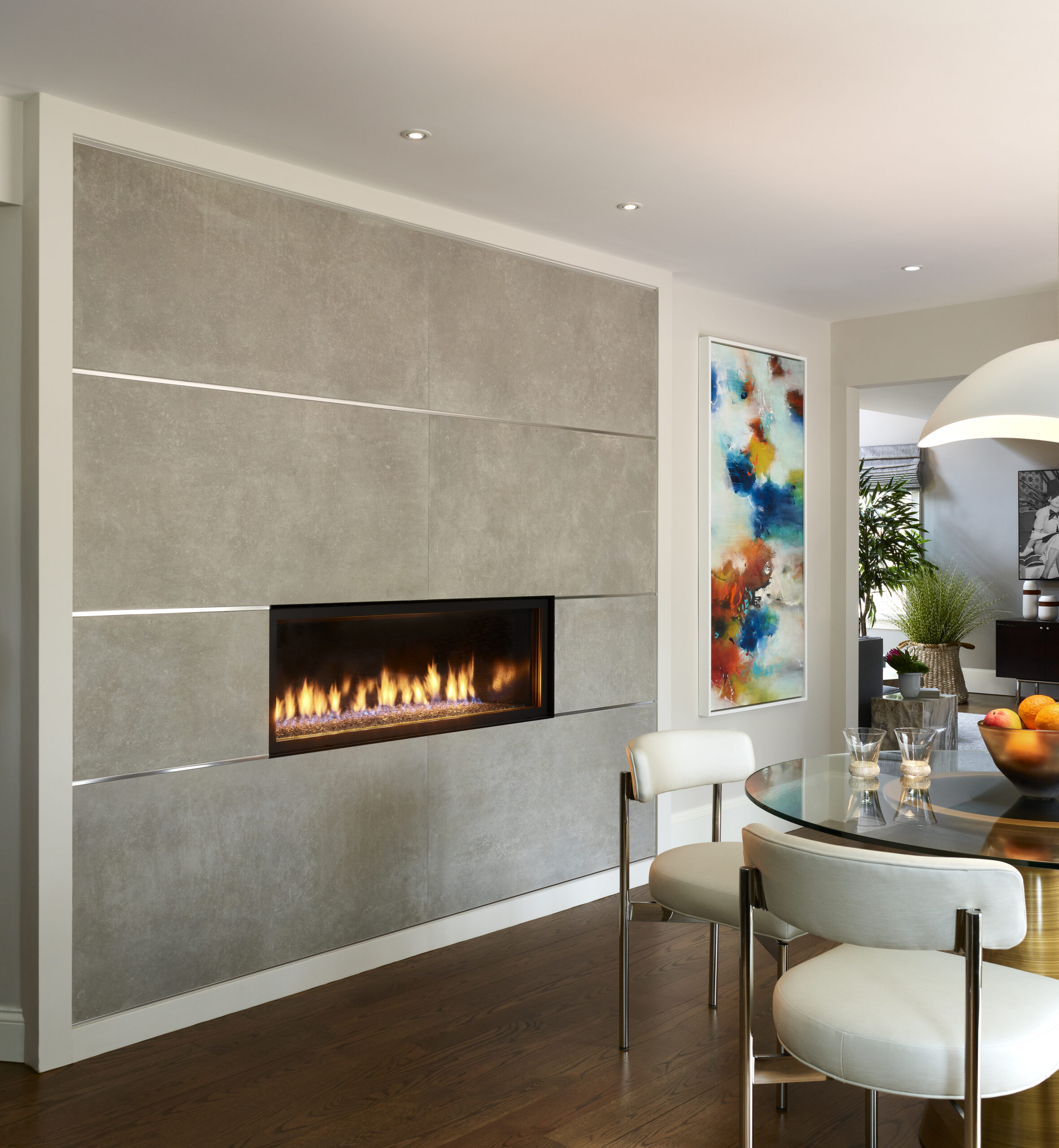 Modern living space with built-in linear fireplace, abstract colorful wall art, round glass dining table, white chairs, and wood flooring—an example of Luxury Interior Design by a top Interior Designer Massachusetts.