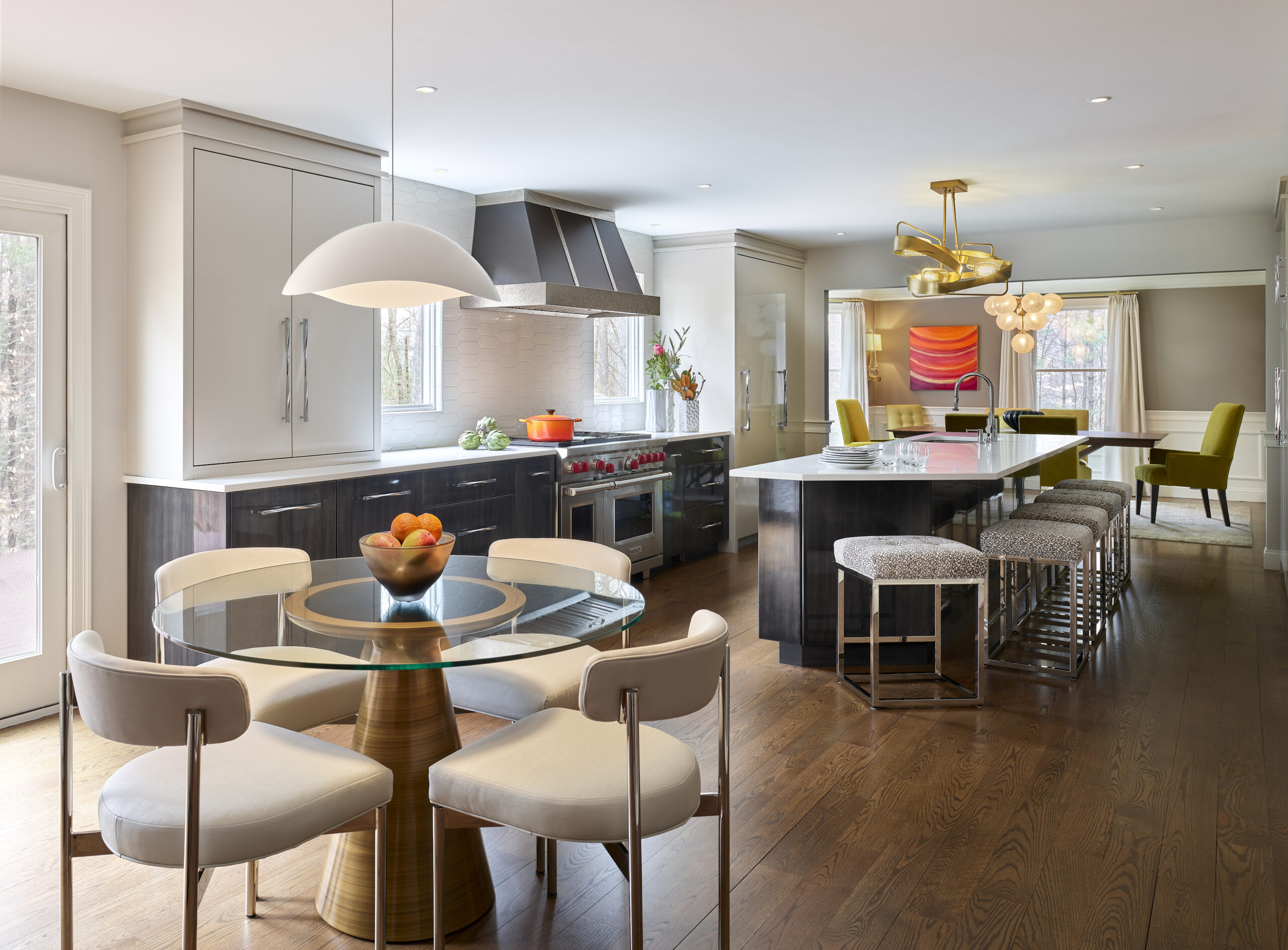 Modern kitchen designed by a top Interior Designer Massachusetts, featuring a round glass dining table, white and gold chairs, large island with stools, stainless steel appliances, and colorful art in the adjacent dining area.