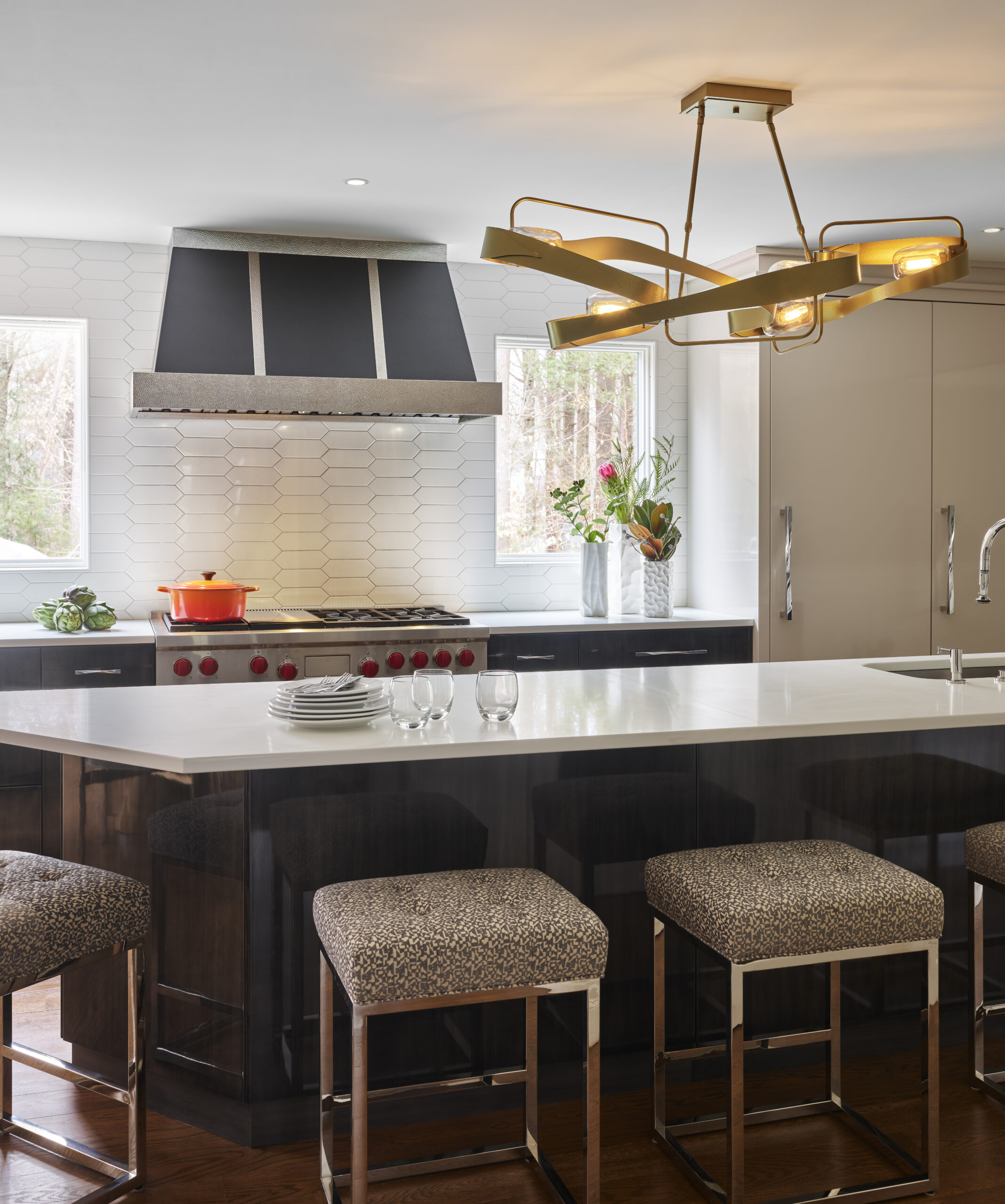 Modern kitchen with a large island, four patterned stools, white countertops, stainless steel appliances, geometric chandelier, and decorative items on the counter—an inspiring example of Luxury Interior Design by an Interior Designer in Massachusetts.