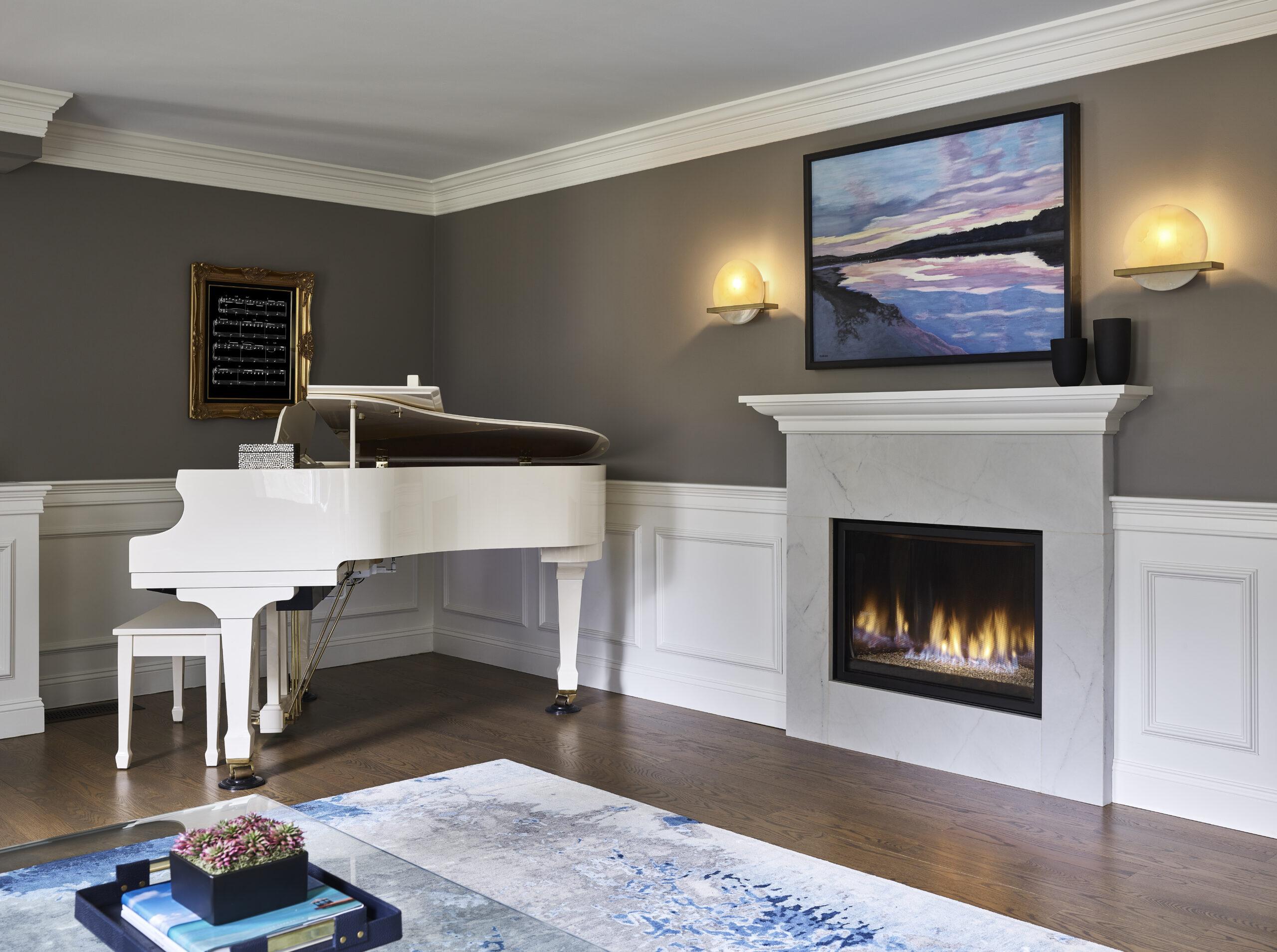 A white grand piano sits near a fireplace with a modern painting above it; this luxury interior design features gray walls, white trim, and a wooden floor with a blue-patterned rug.