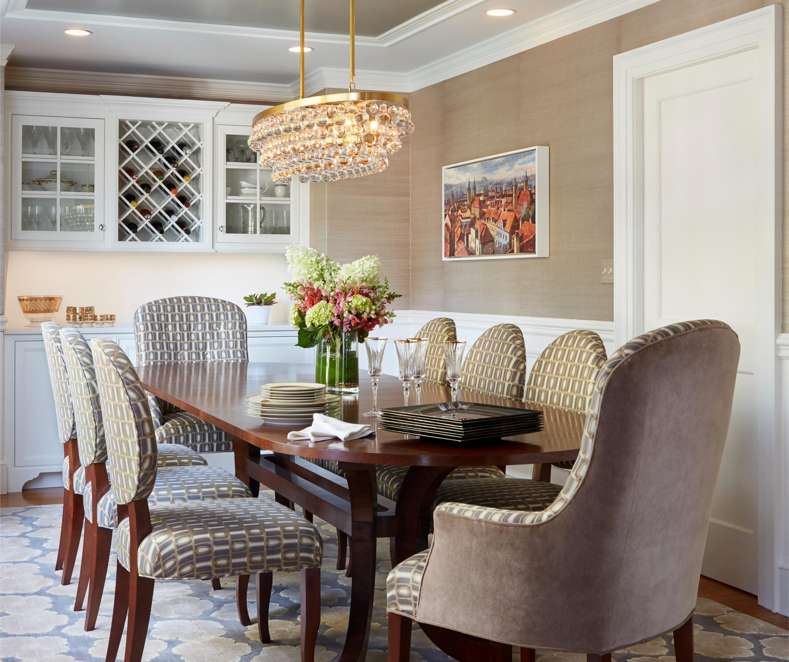 A dining room with a wooden table, patterned chairs, a crystal chandelier, flowers in vases, plates, glasses, and cabinetry with a wine rack along one wall—expertly styled through professional Interior Design Services.