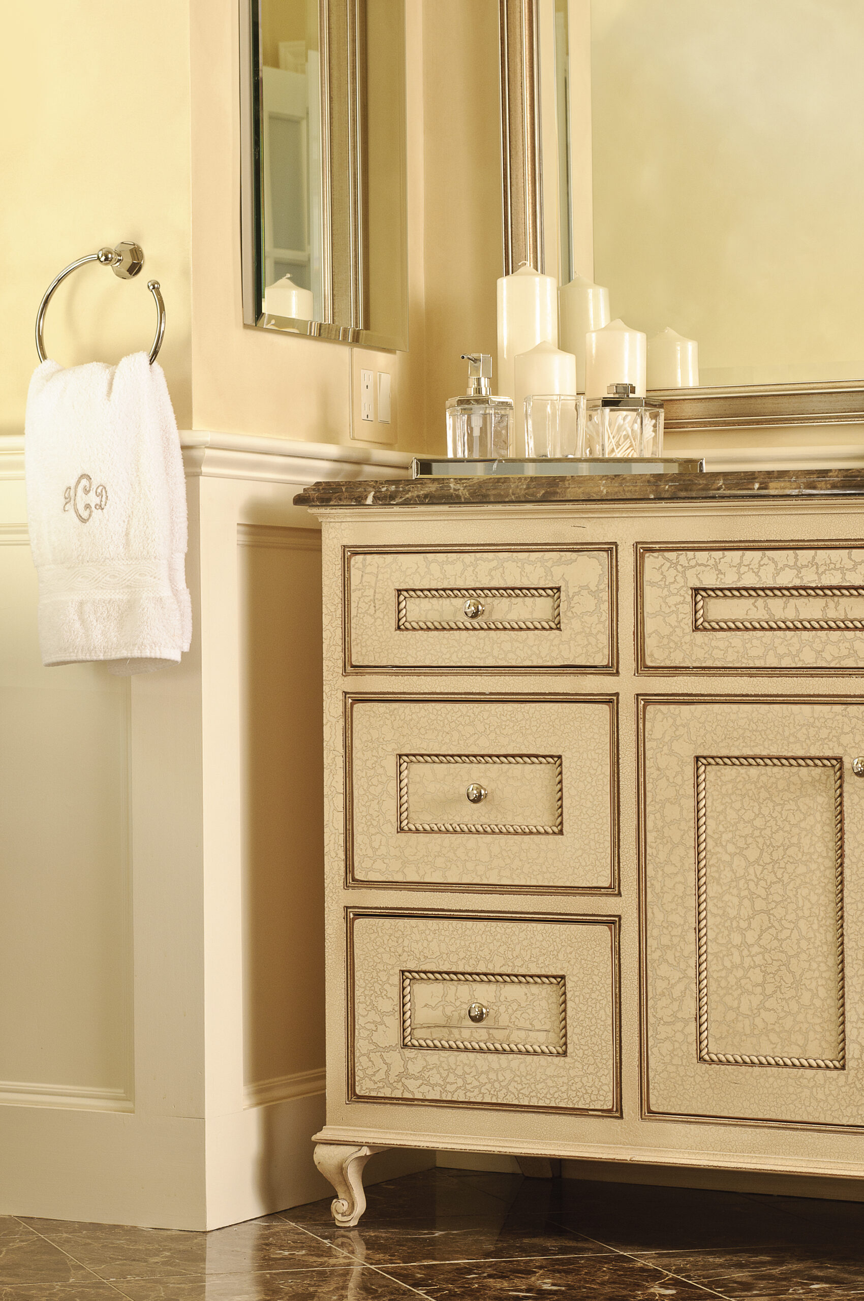 Beige bathroom vanity with a marble countertop, decorative drawers, glass containers, and white candles beside a towel hanging on a metal ring—perfect for elevating your space with Luxury Interior Design.