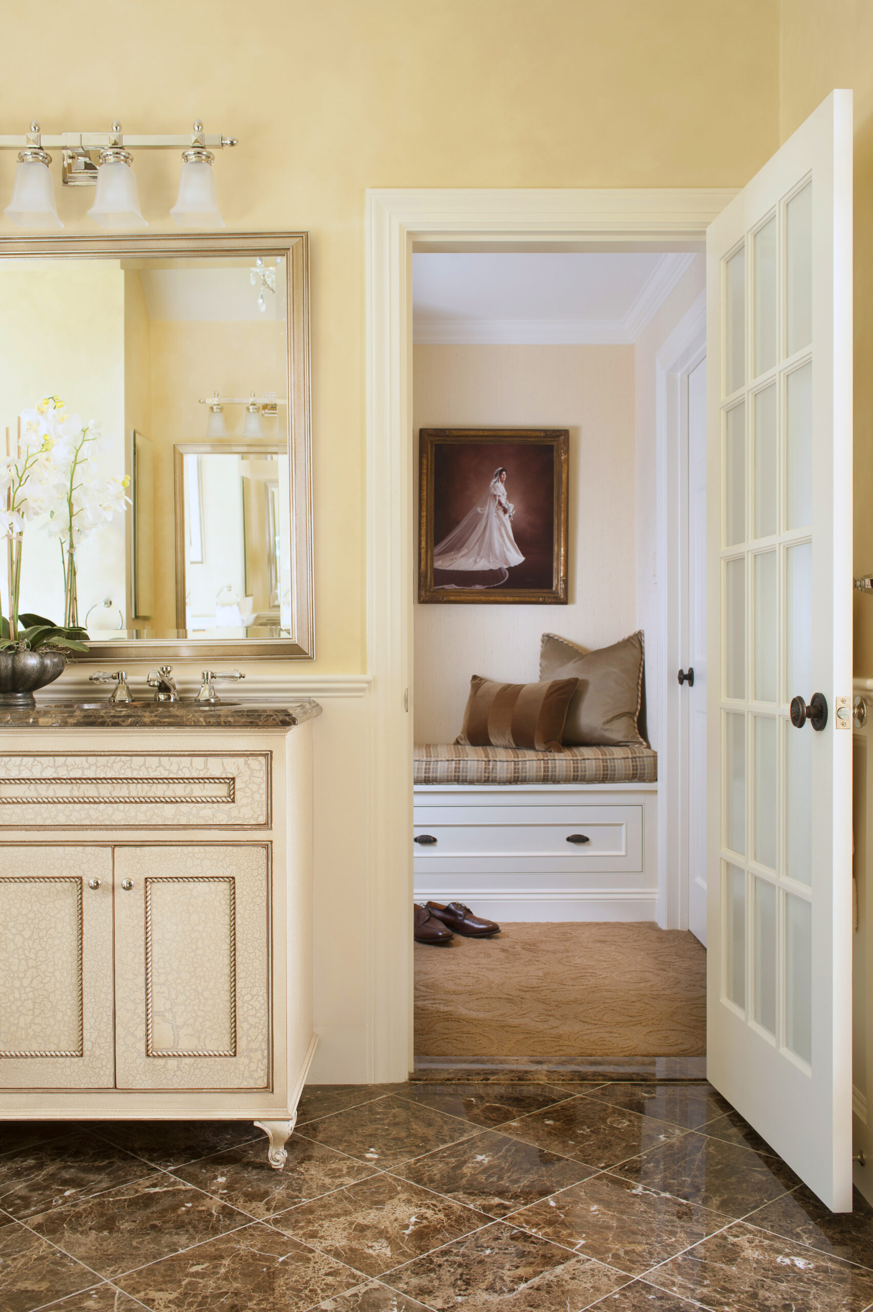 A bathroom with a beige vanity and marble floor opens to a room with a cushioned bench, throw pillows, and a framed portrait—an elegant touch of luxury interior design that echoes the style of leading Massachusetts designers. Shoes rest on the carpet nearby.