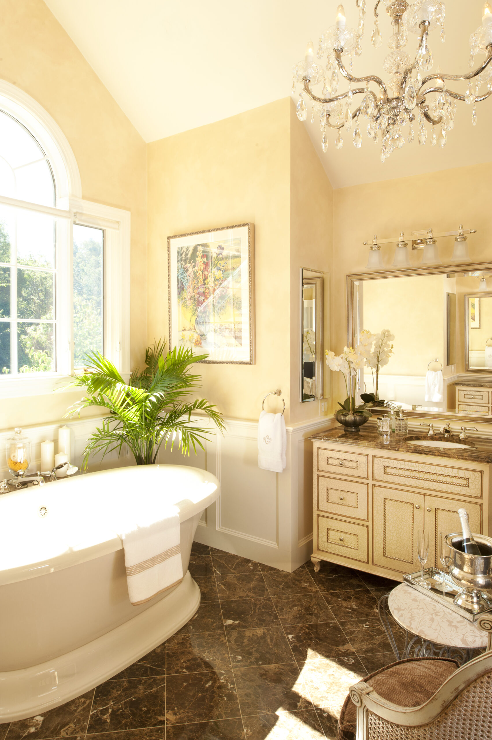 Elegant bathroom with a freestanding tub, marble floor, chandelier, and large window—showcasing Luxury Interior Design at its finest. A potted plant and a vanity with mirror and flowers complete this sophisticated retreat.