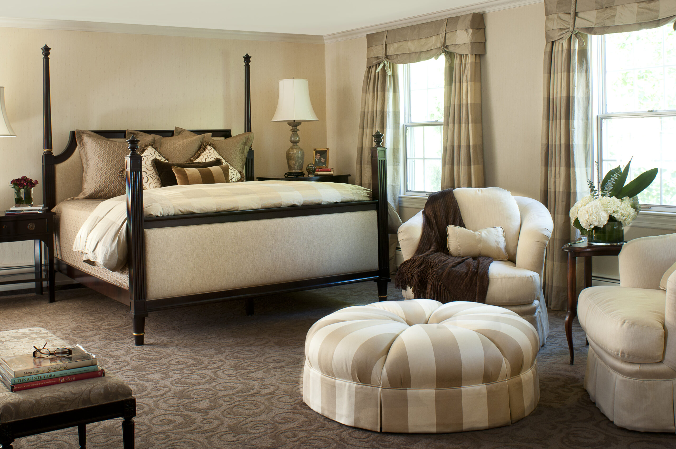 A well-lit bedroom by a top Interior Designer Massachusetts, featuring a four-poster bed, two armchairs, a round striped ottoman, side tables with lamps, and large windows with beige curtains for a touch of luxury interior design.