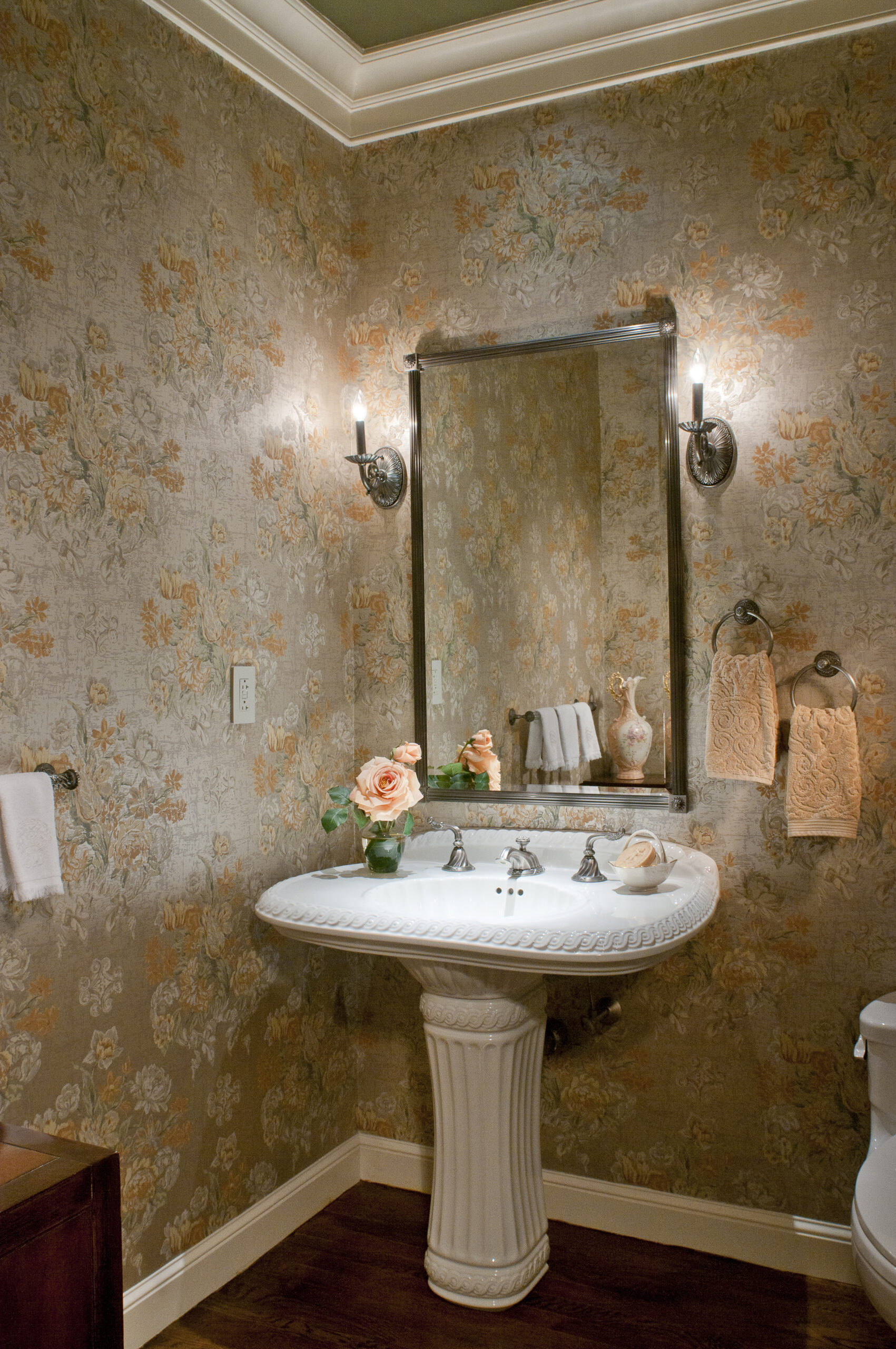 A vintage-style bathroom by a top Interior Designer Massachusetts features floral wallpaper, a pedestal sink, a rectangular mirror, wall sconces, and decorative peach roses for a touch of luxury interior design charm.