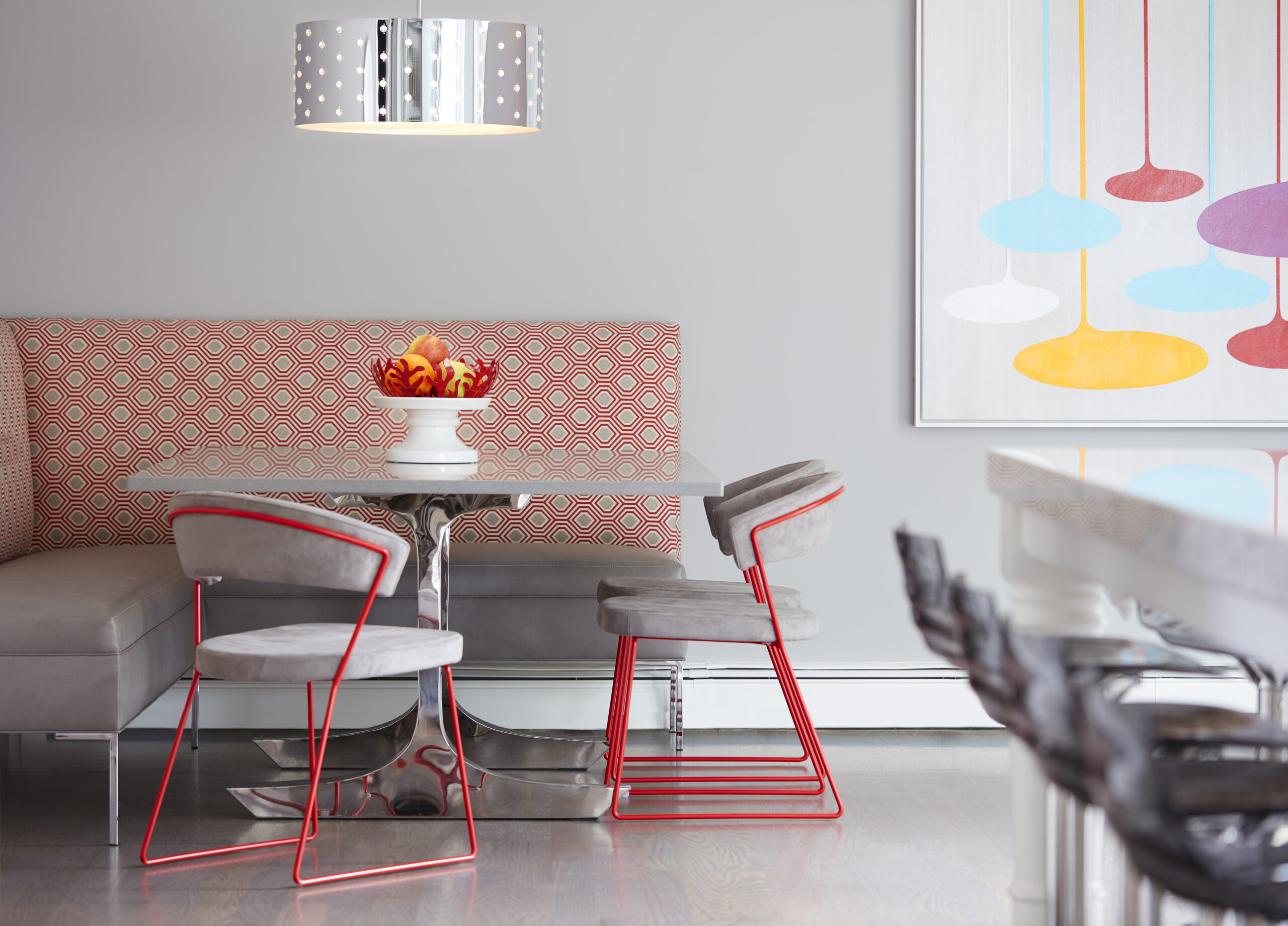 Modern dining area with a patterned booth, gray chairs with red legs, a round table, a bowl of flowers, and a colorful abstract art piece on a gray wall—an example of luxury interior design by an expert interior designer in Massachusetts.