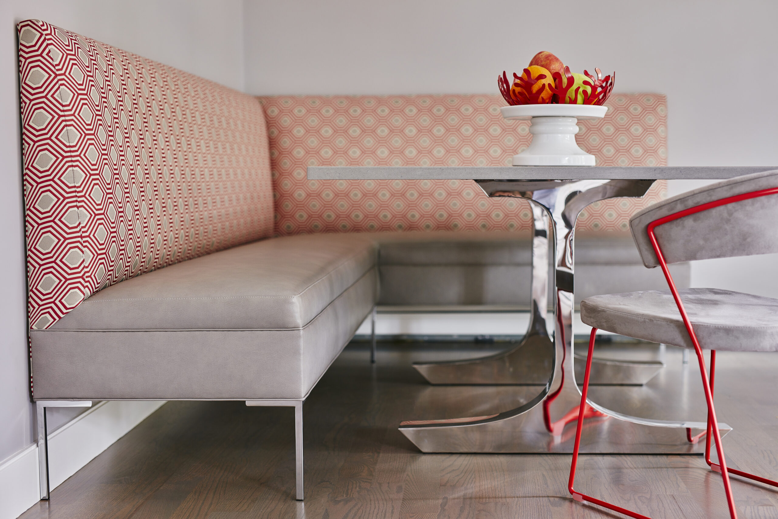 Modern dining nook with a patterned upholstered bench, chrome table, gray cushion, and red-accented chairs; a white bowl of fruit sits on the table—showcasing luxury interior design worthy of any top interior designer in Massachusetts.