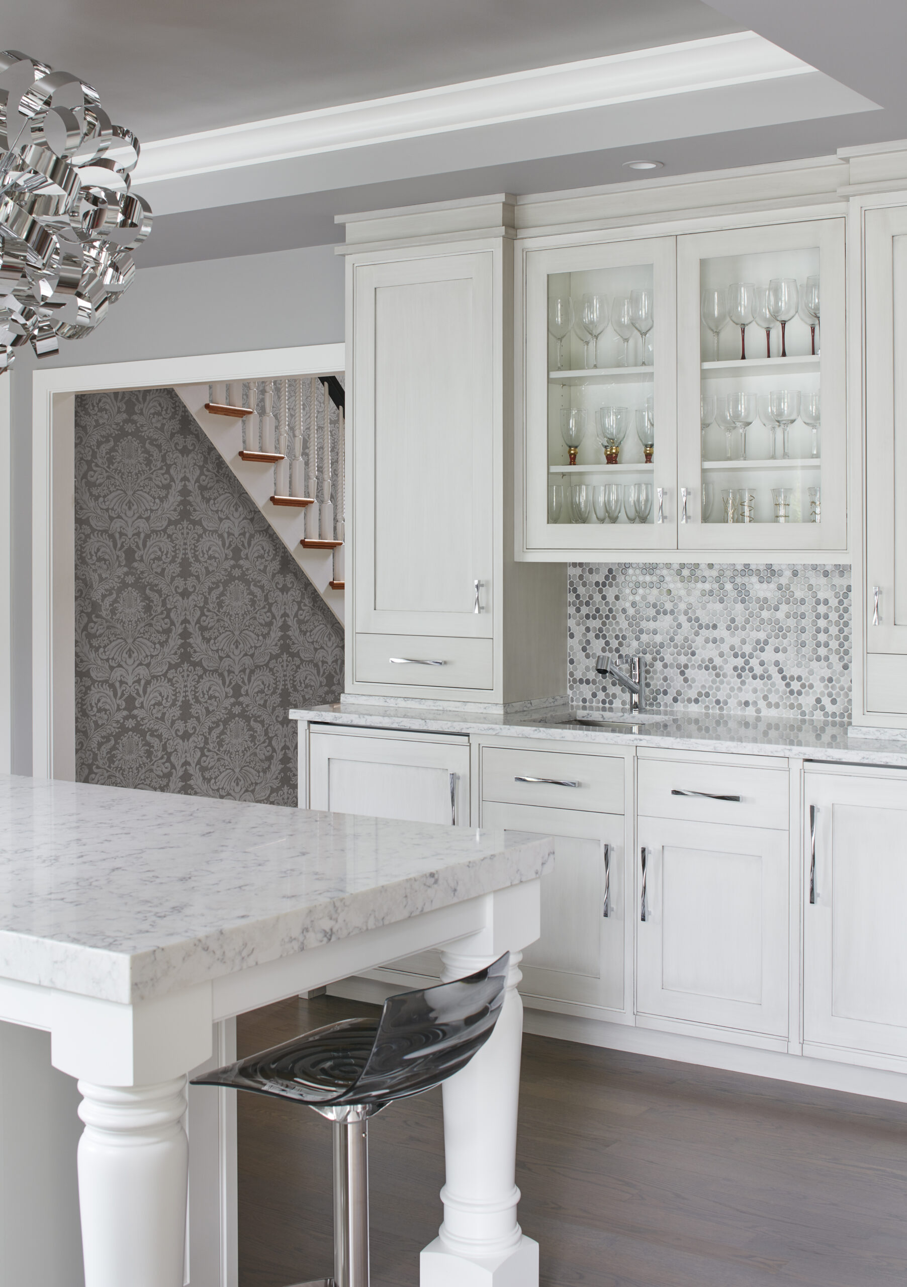 Modern white kitchen with marble countertops, glass-front cabinets displaying glassware, mosaic tile backsplash, and a clear acrylic barstool. A showcase of luxury interior design by a leading interior designer in Massachusetts; staircase visible in the background.