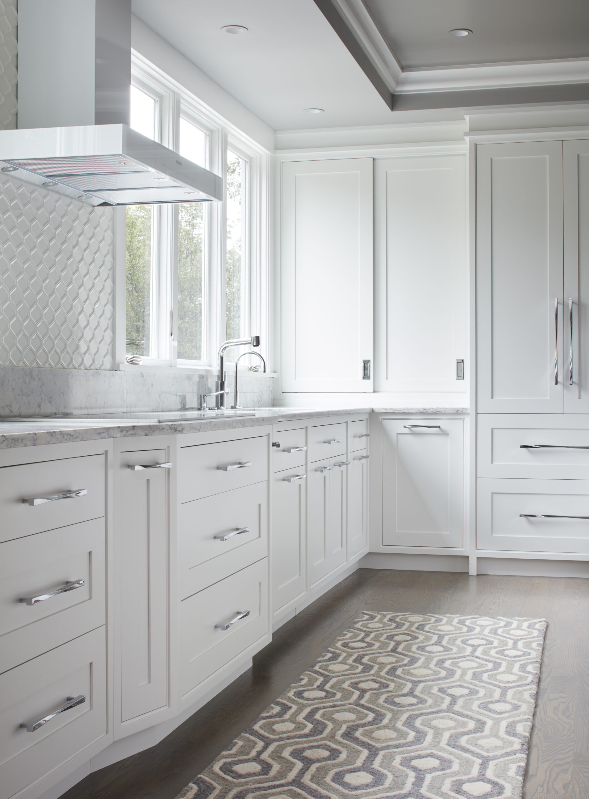 Modern kitchen with white cabinets, marble countertops, stainless steel handles, large windows, and a geometric-patterned rug on a wooden floor—an elegant space crafted by an expert in luxury interior design.