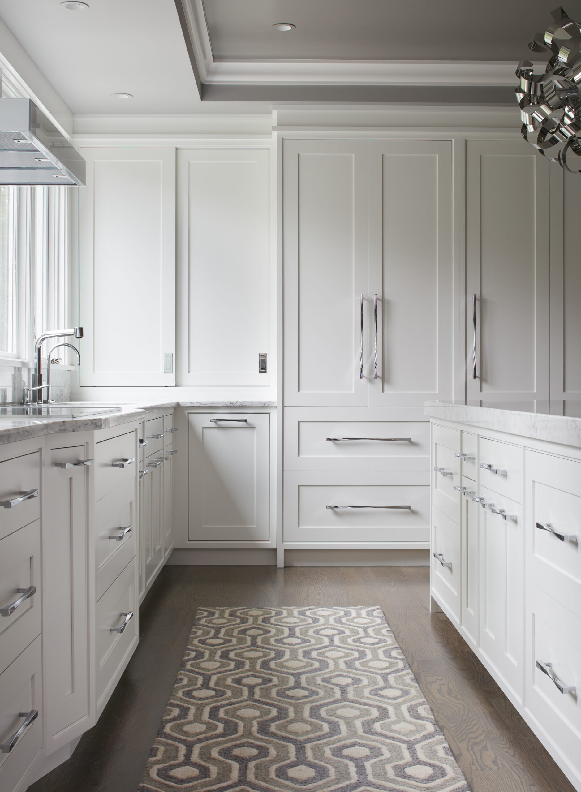 Modern kitchen with white cabinets, stainless steel handles, marble countertops, and a patterned rug on a wooden floor. Large windows provide natural light—an example of luxury interior design by an expert interior designer in Massachusetts.