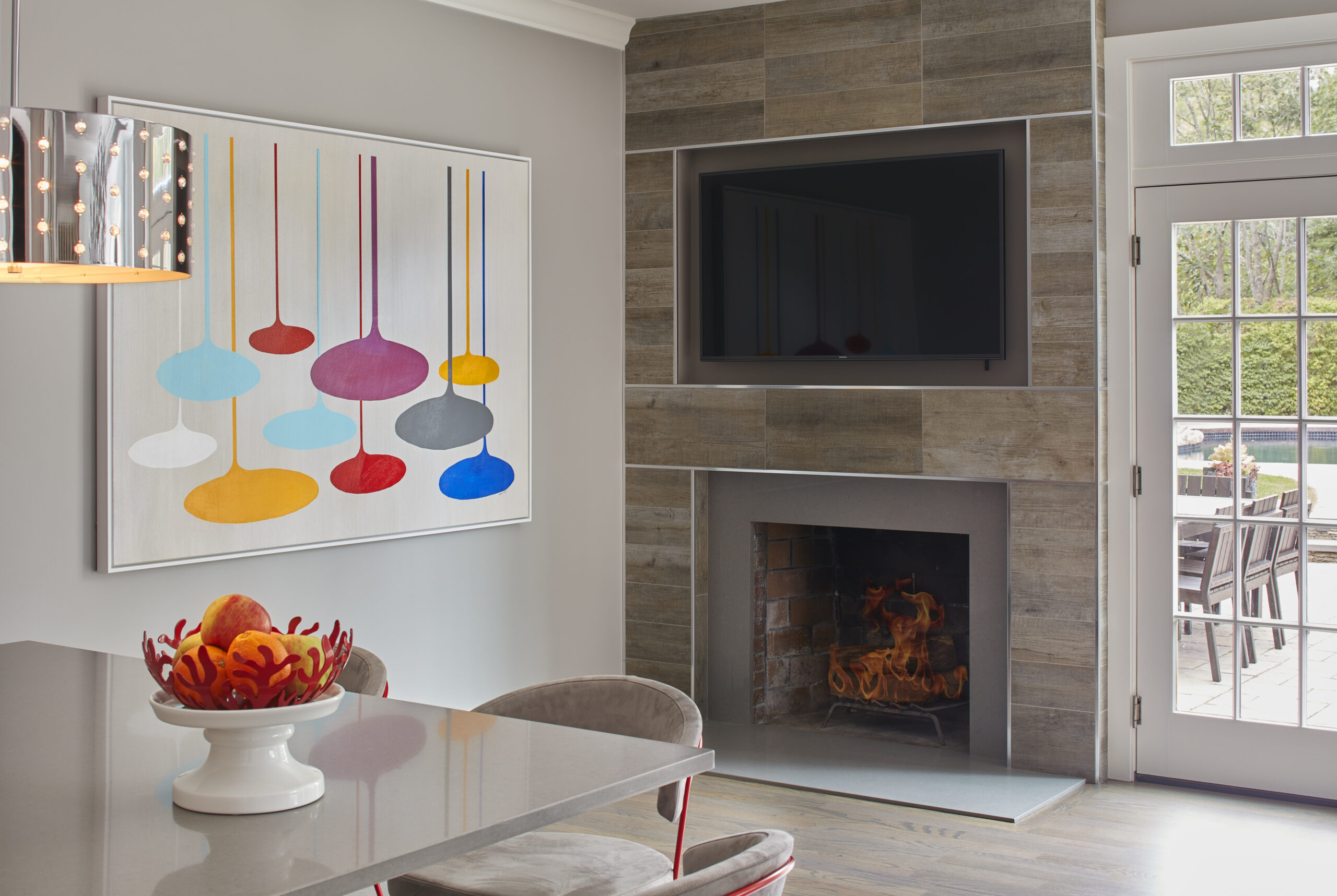 Modern living area with a wall-mounted TV above a fireplace, colorful abstract art, and a dining table with a fruit bowl centerpiece—showcasing Luxury Interior Design by an expert interior designer in Massachusetts.