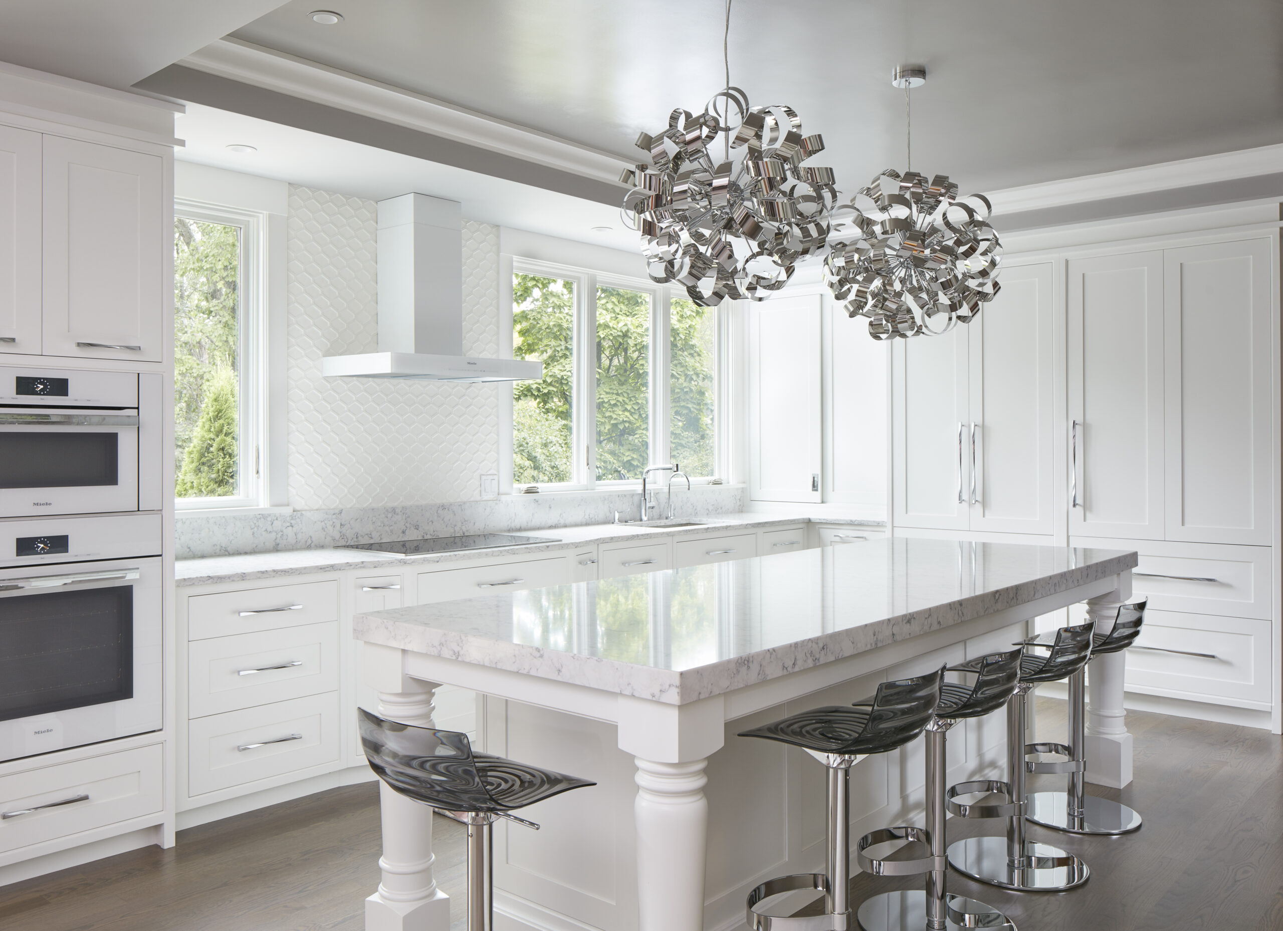 Modern white kitchen with marble island, chrome bar stools, built-in appliances, large windows, and sculptural pendant lights—perfectly crafted by an expert Interior Designer Massachusetts for luxury interior design enthusiasts.