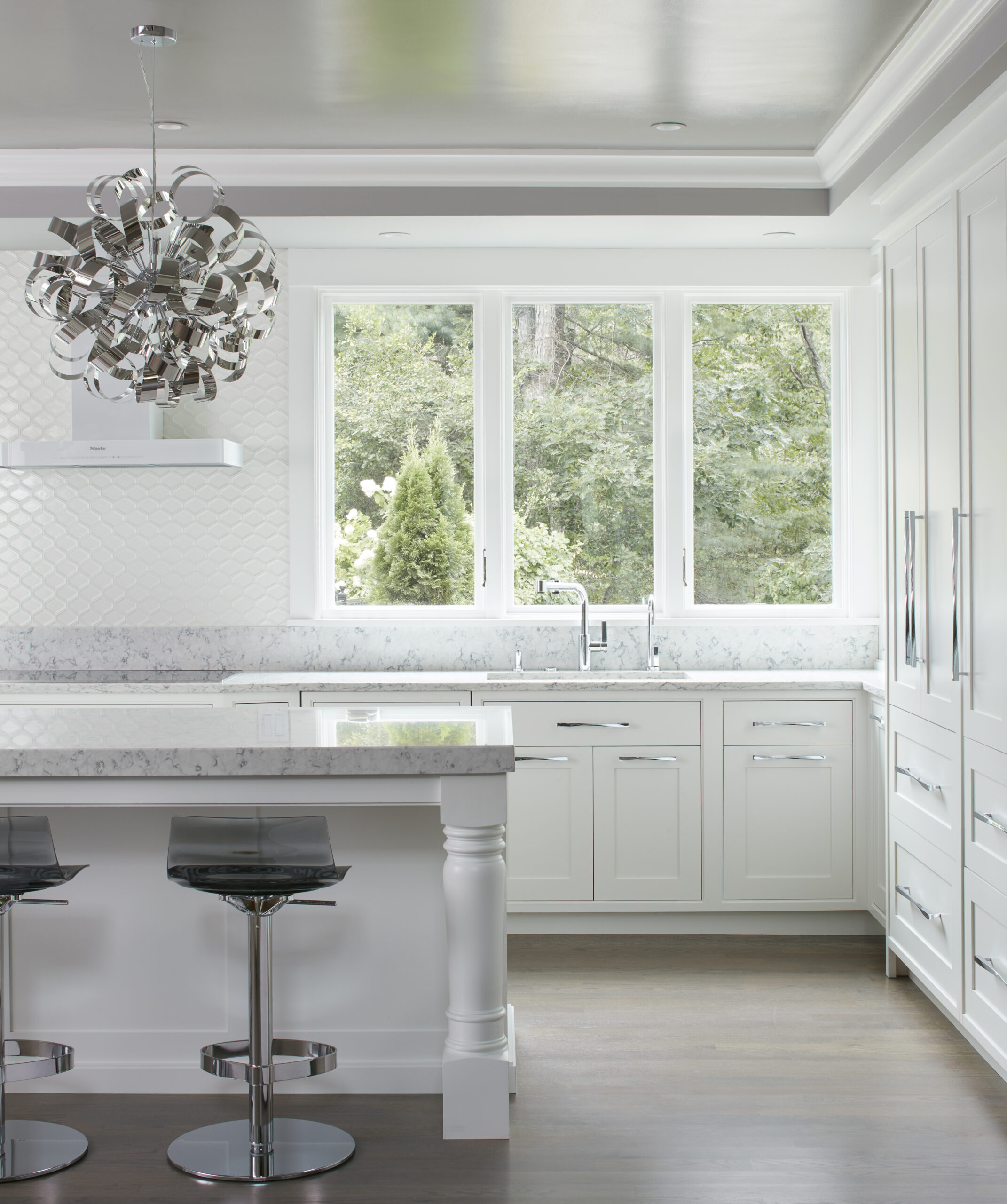 Modern white kitchen with marble countertops, a large window overlooking greenery, high-gloss bar stools, and a metallic contemporary chandelier—showcasing refined Luxury Interior Design by a skilled Interior Designer Massachusetts.