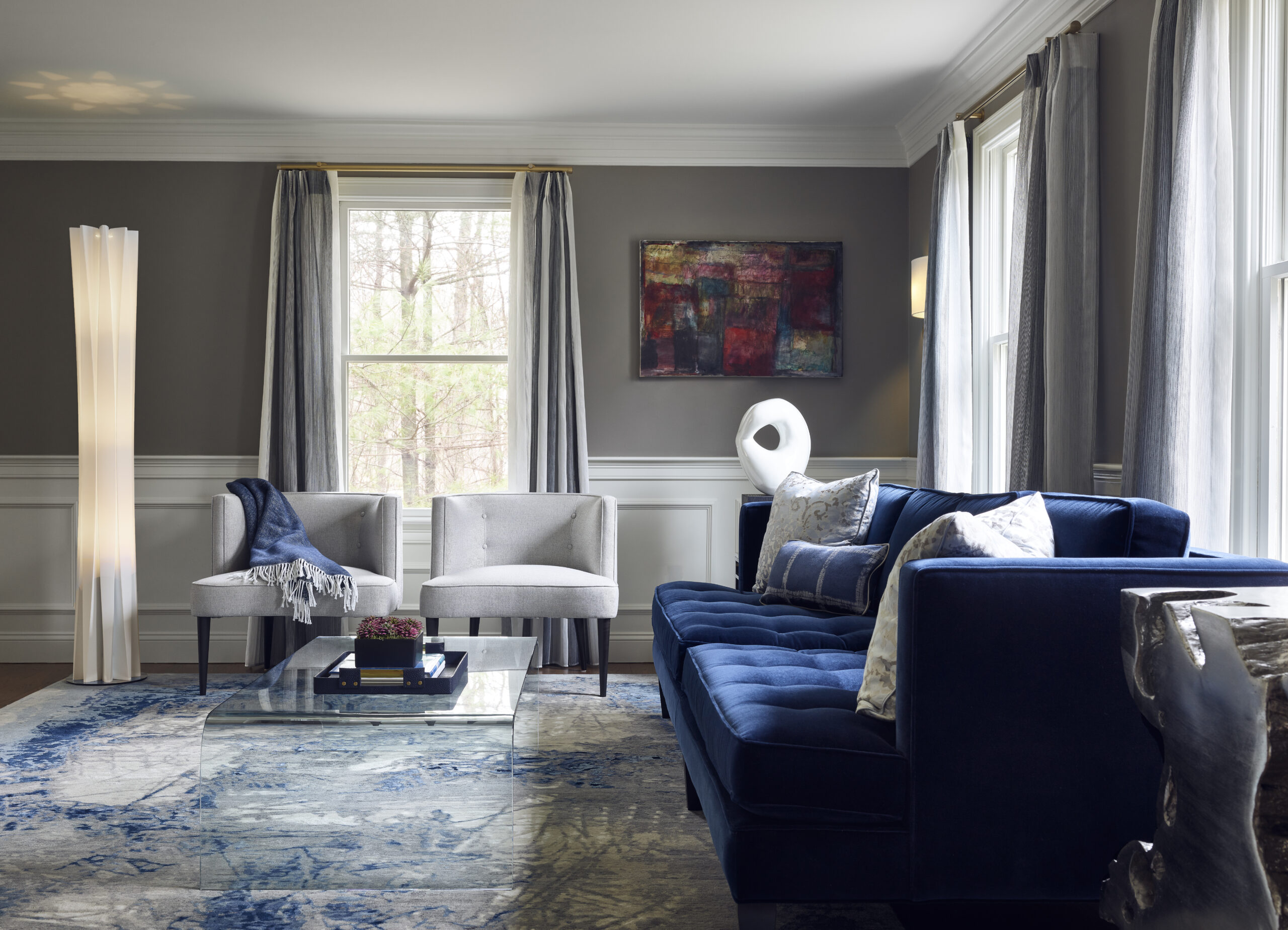 A modern living room by an Interior Designer Massachusetts features a blue velvet sofa, two gray chairs, a glass coffee table, abstract art on the wall, and large windows with gray curtains for true luxury interior design.