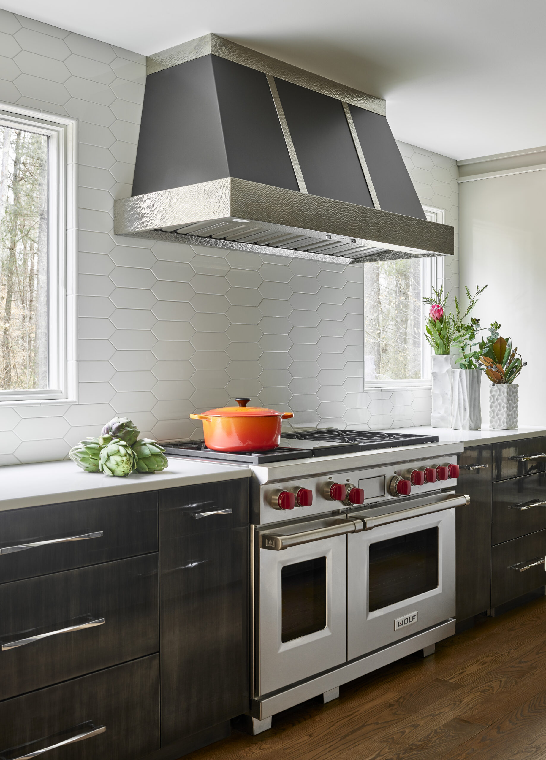Modern kitchen with stainless steel stove and range hood, orange pot on stovetop, white hexagonal tile backsplash, and vases with flowers on the counter—perfectly reflecting luxury interior design by a top Interior Designer in Massachusetts.