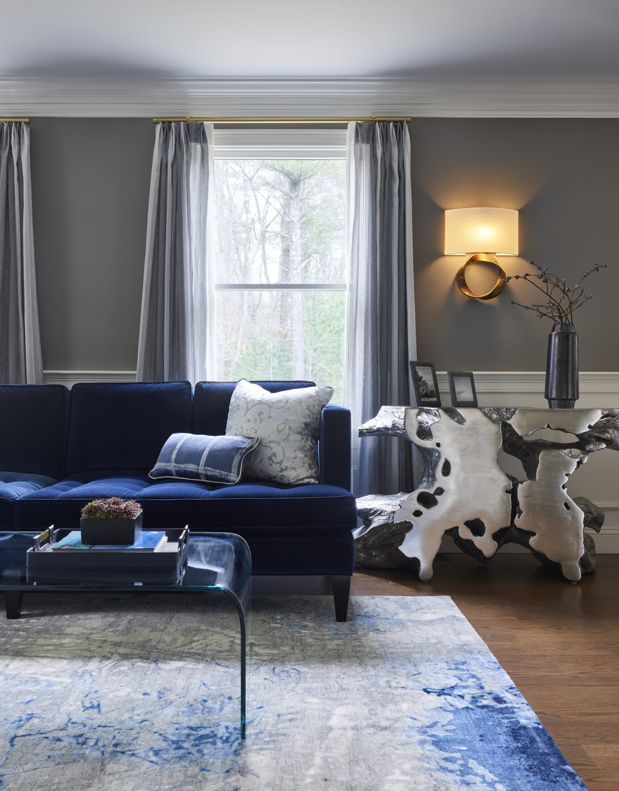 Modern living room with a navy velvet sofa, patterned pillows, abstract wood side table, glass coffee table, and gray curtains—an inviting space that reflects luxury interior design by an expert Interior Designer Massachusetts.