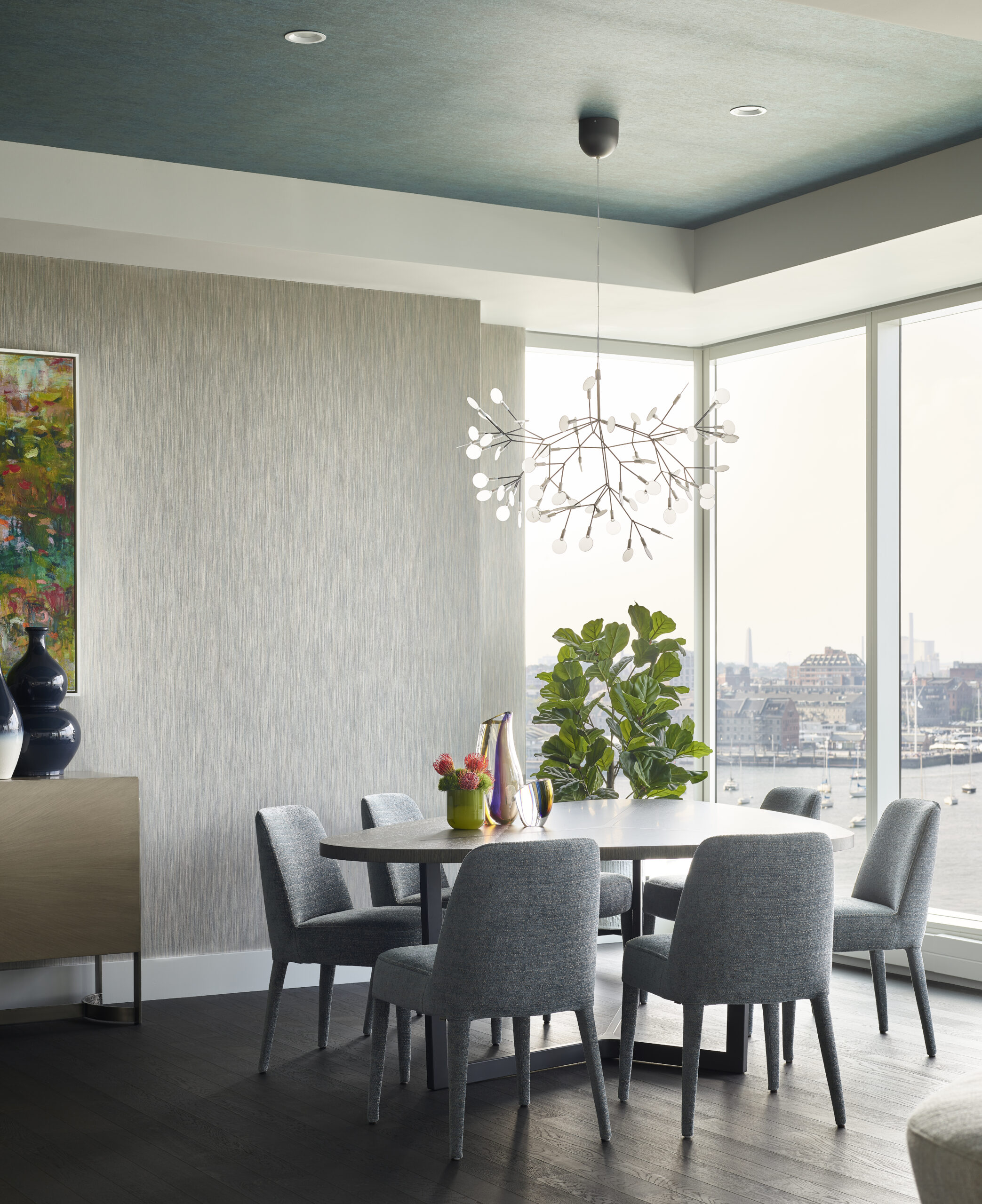Modern dining room with six gray chairs around a table, a contemporary chandelier, potted plant, large window with city view, and abstract art highlights luxury interior design by a top Interior Designer Massachusetts.