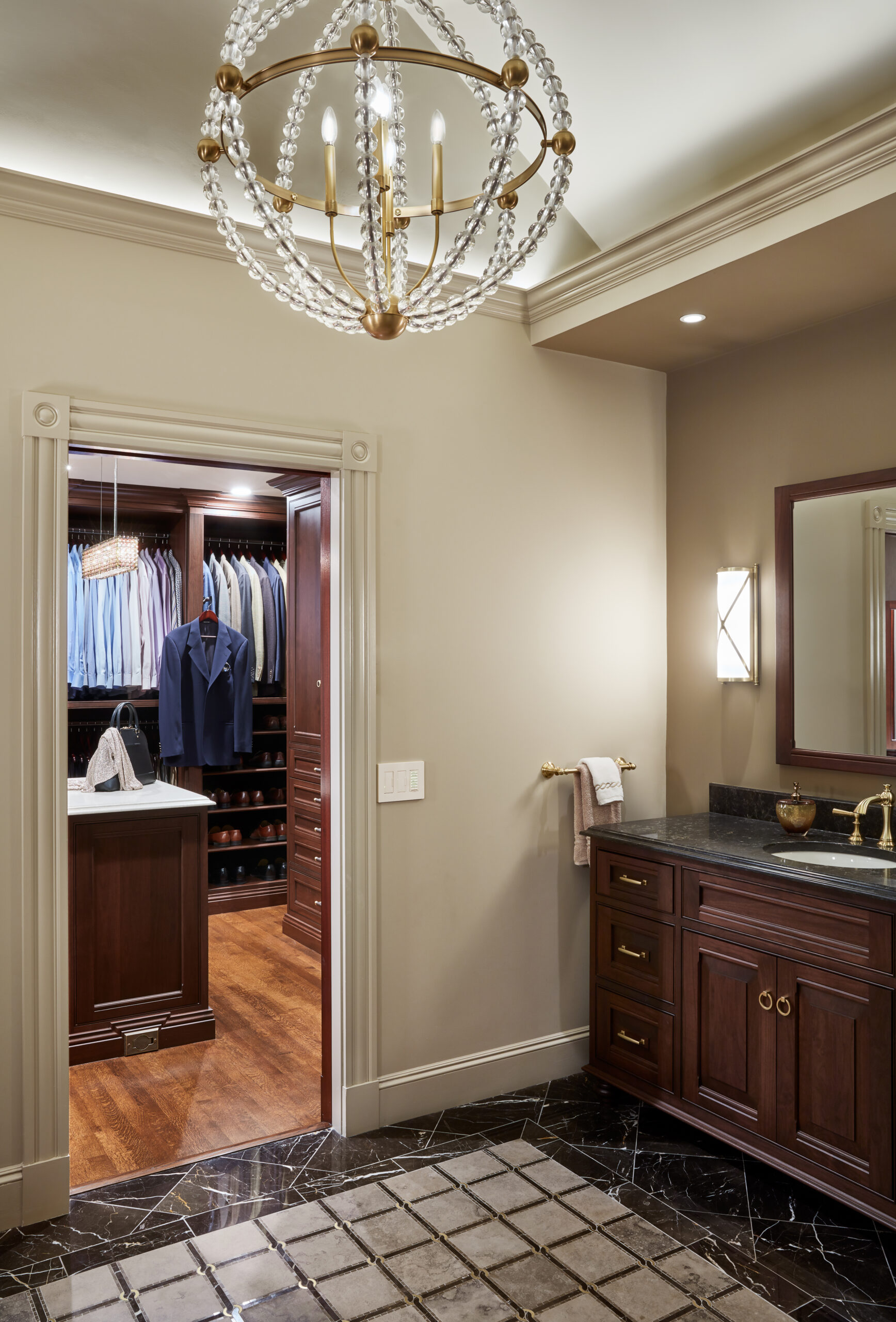 A bathroom with a dark marble floor and wood vanity, designed with luxury interior design in mind, leads to a walk-in closet with hanging clothes, drawers, and an island; a chandelier hangs from the ceiling.