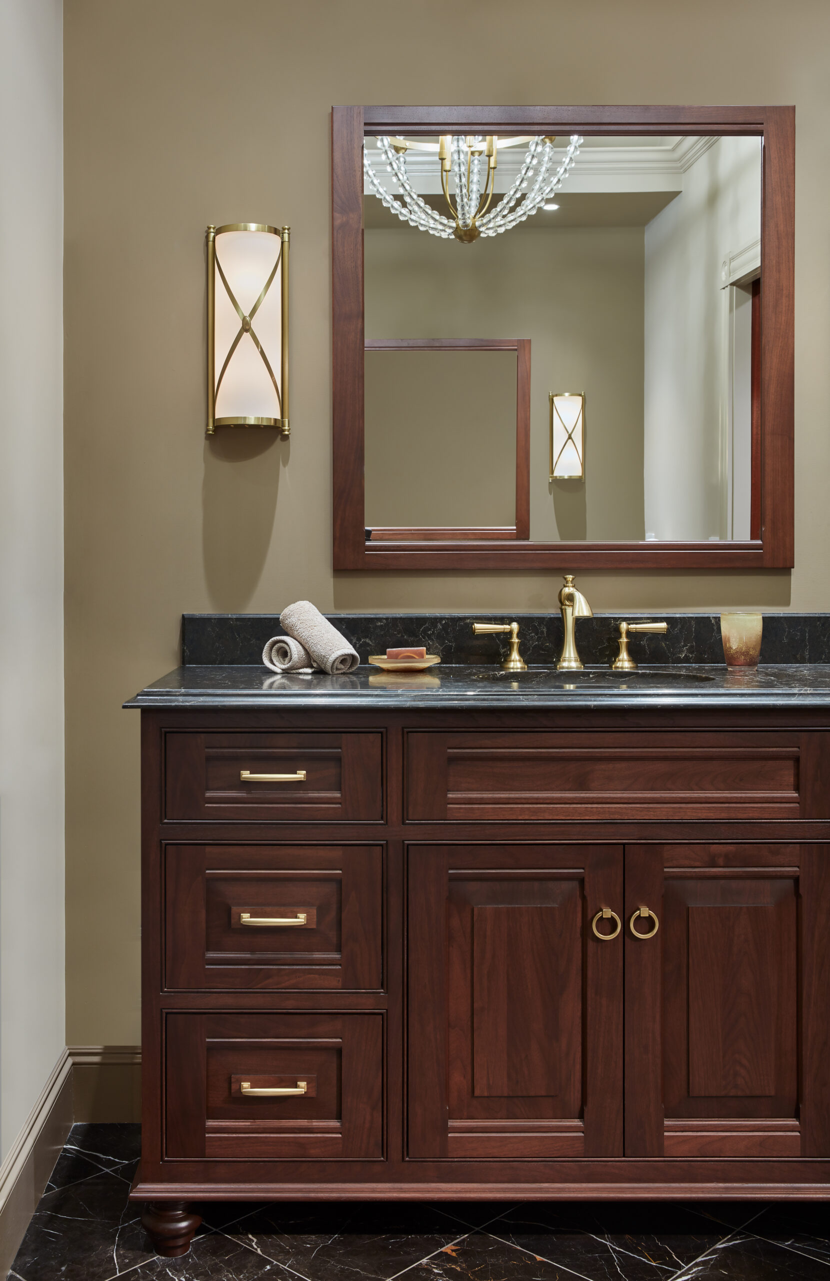 A bathroom vanity with dark wood cabinets, black countertop, gold fixtures, a mirror, wall sconce, and two rolled towels showcases luxury interior design perfect for those seeking elegance in their Massachusetts home.