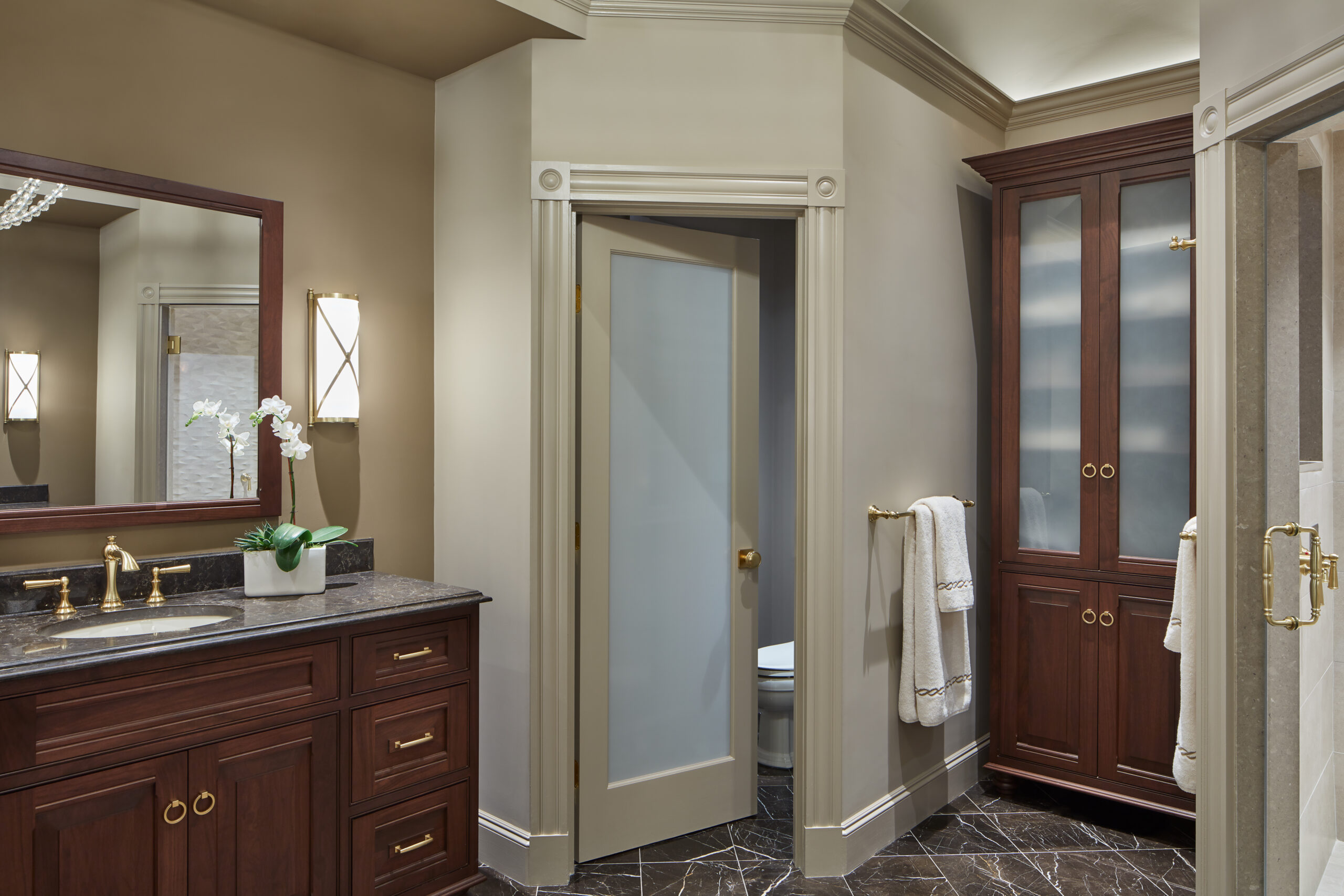 Traditional bathroom with dark wood cabinets, marble countertop and floor, frosted glass door, and brass fixtures. Designed by an Interior Designer in Massachusetts, this luxury interior design features towels hanging beside a tall cabinet.